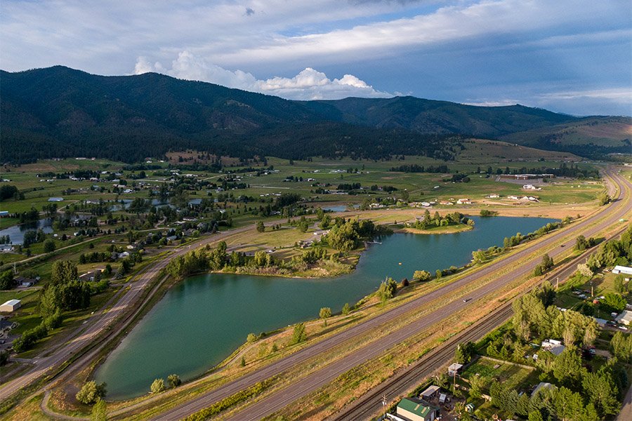 A scenic aerial view of a small town with a lake, lush greenery, and a mountainous backdrop under a partly cloudy sky.