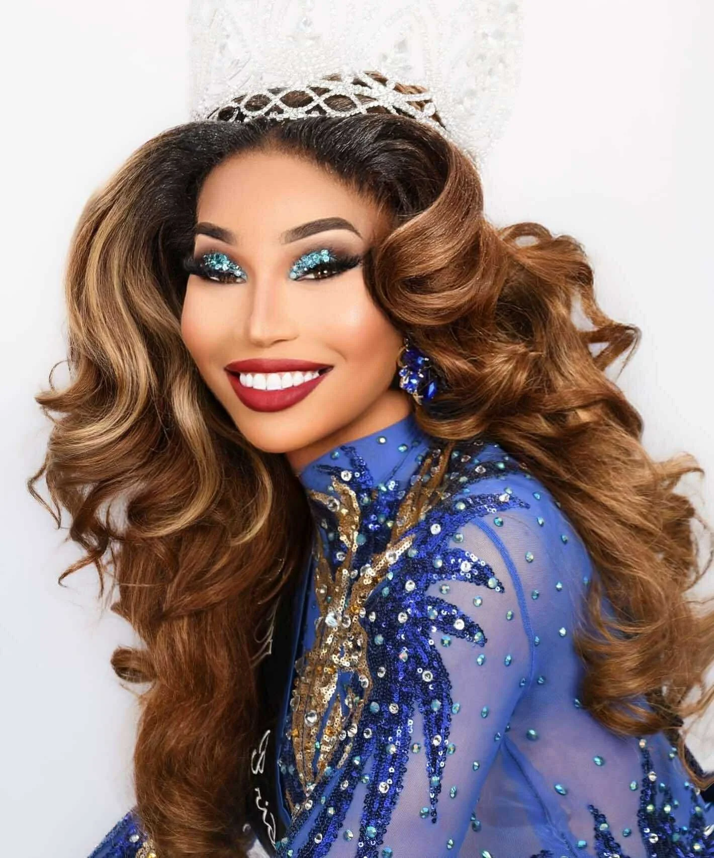 A woman with long, styled brown hair, wearing a blue dress with gold and blue embellishments, and a silver crown. She has makeup with glittery blue eyeshadow and red lipstick, and is smiling.