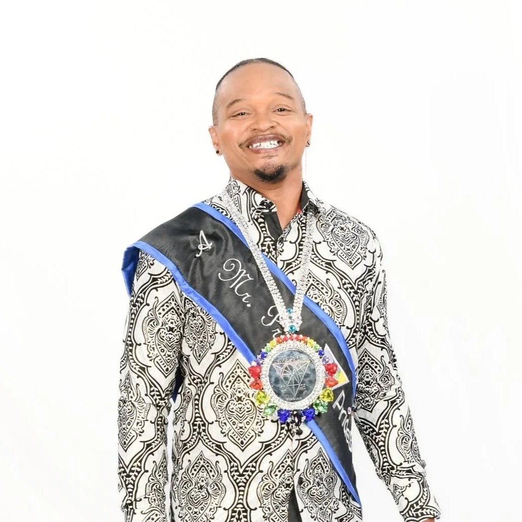 A smiling man wearing a black and white patterned shirt and a sash with 'Mr. K' on it, adorned with a large necklace with a colorful pendant, standing against a plain white background.