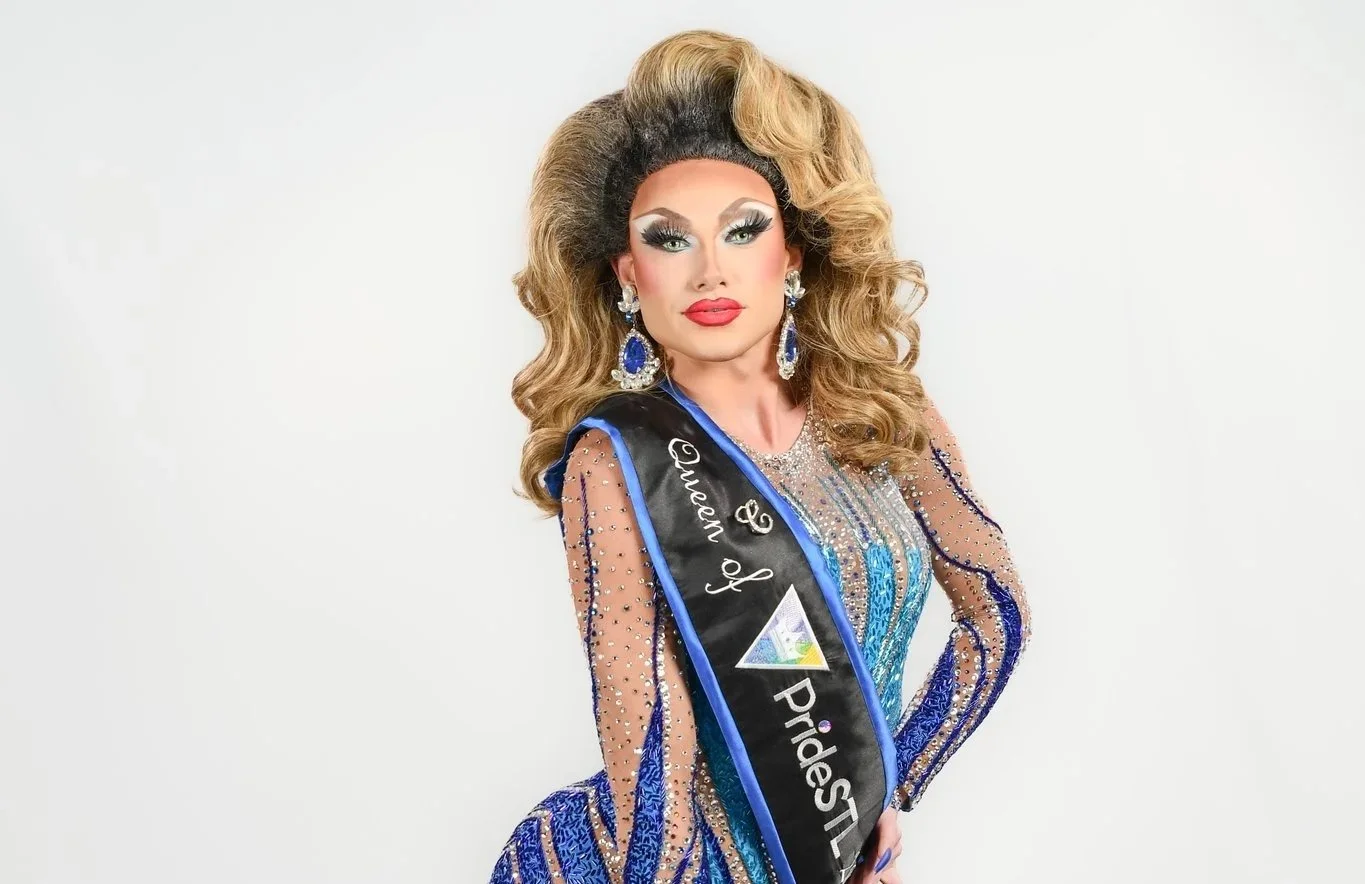 A drag queen with voluminous blonde hair styled with dark roots, wearing large blue earrings, a blue and black sash reading "Queen of Pride" over a sparkly, sheer, blue and gold dress with rhinestone embellishments, posing with one hand on her hip against a plain white background.