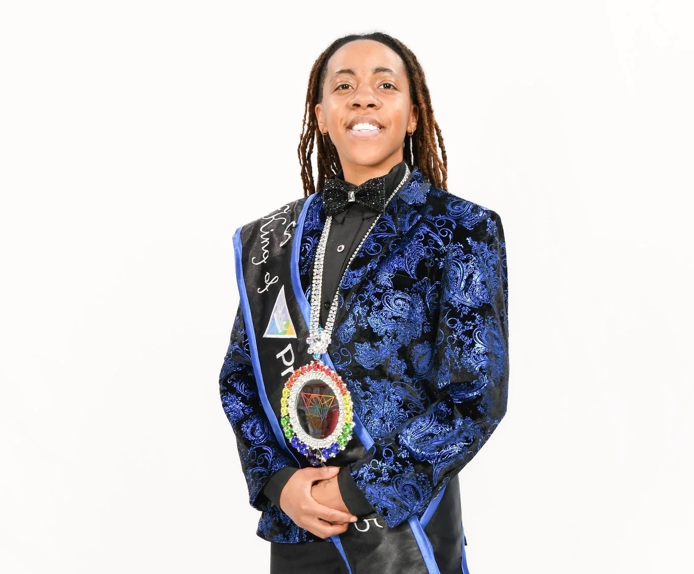 Individual wearing a blue velvet tuxedo jacket with black shirt, black bow tie, standing against white background, with a sash and large medallion, smiling.