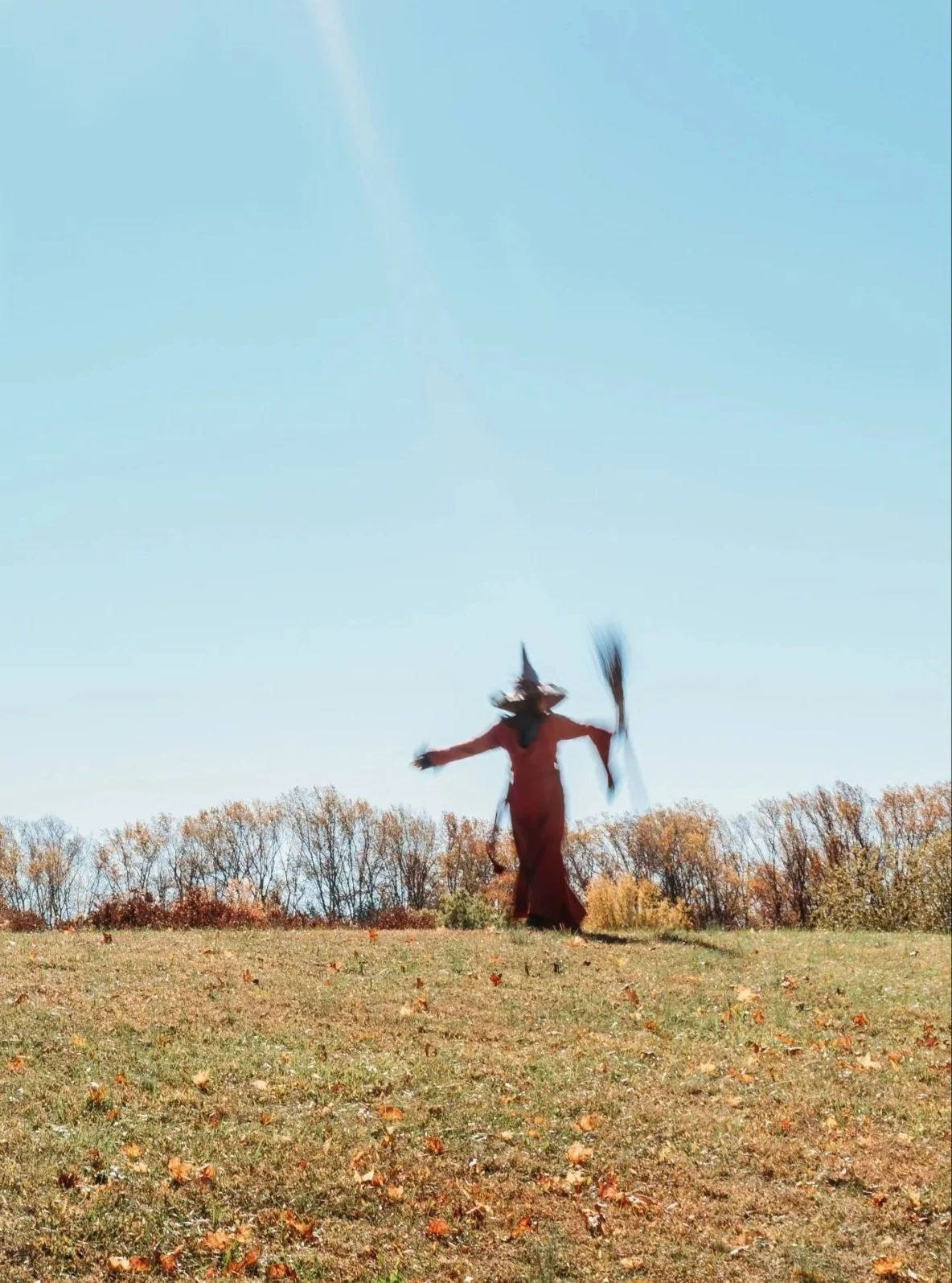 Woman in a witch outfit walking up a hill in a field. She is wearing a purple hat, red cape, and is carrying a broom.