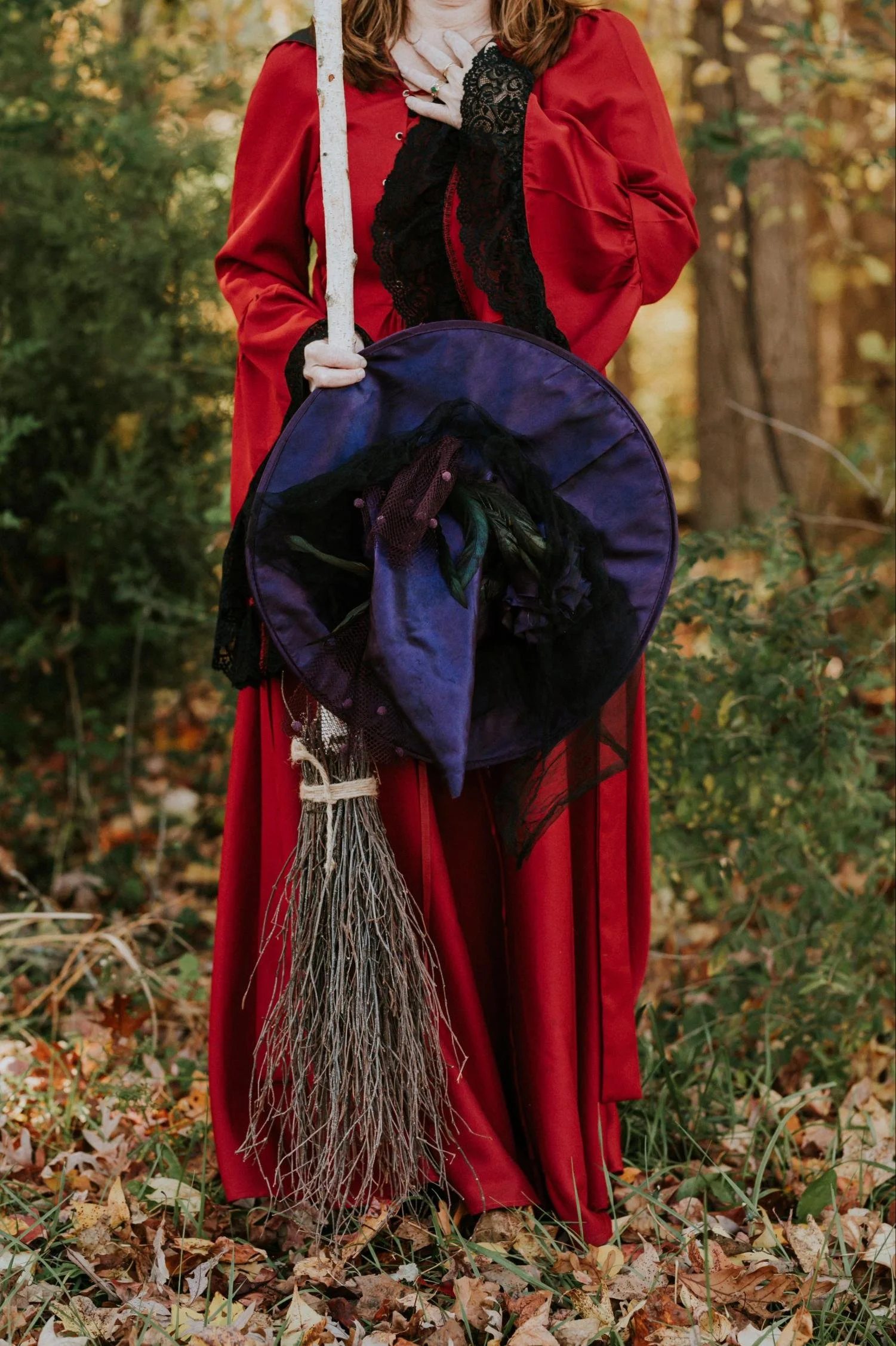 Close up of a witch outfit, including a woman standing in grass and leaves wearing a red cape, purple witch hat, and holding a broom made of branches and tied with woven string.