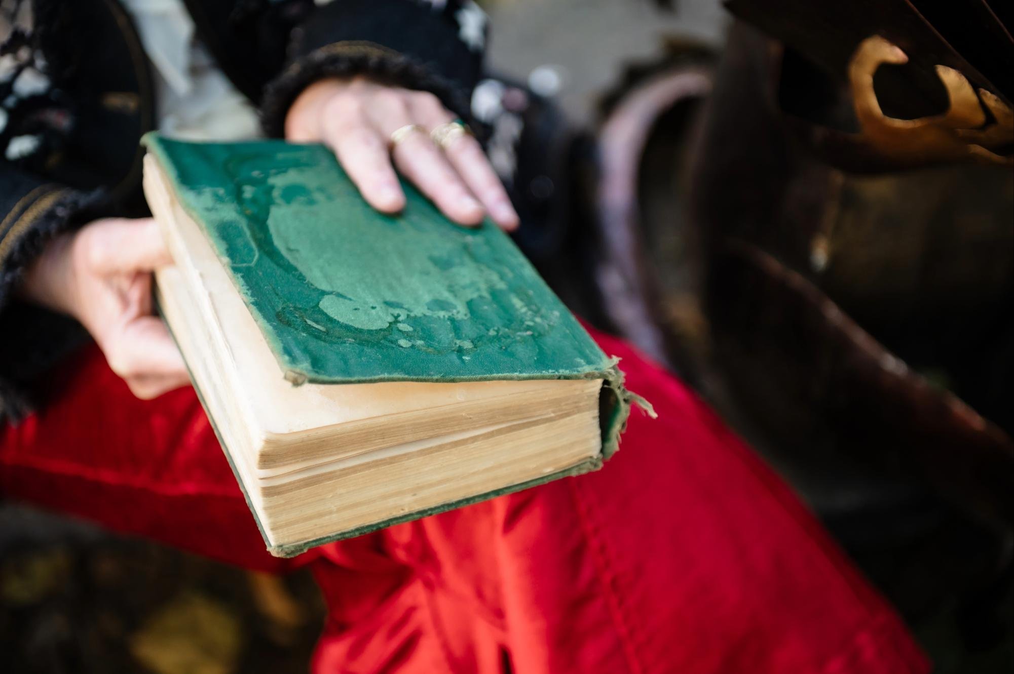 Woman holding a vintage book while sitting outside. Close up of the book, which has aged paper. The binding is coming apart, and there is natural wear on the turquoise cover.