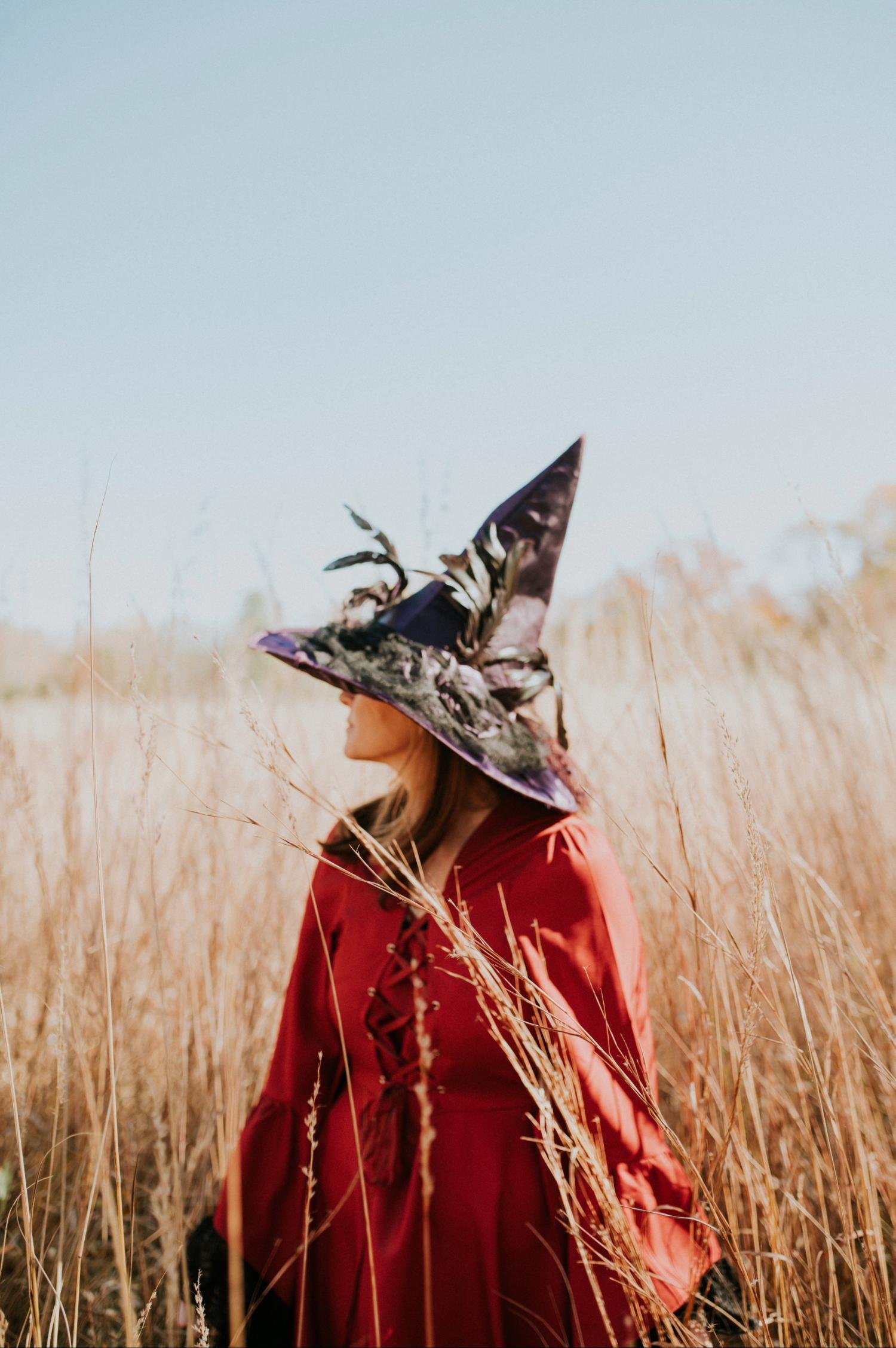 Woman in a witch outfit standing in a field. She's wearing a purple witch hat and a red cape, and this image is being used to represent Witches of Eastport, a service provider offering astrology services.