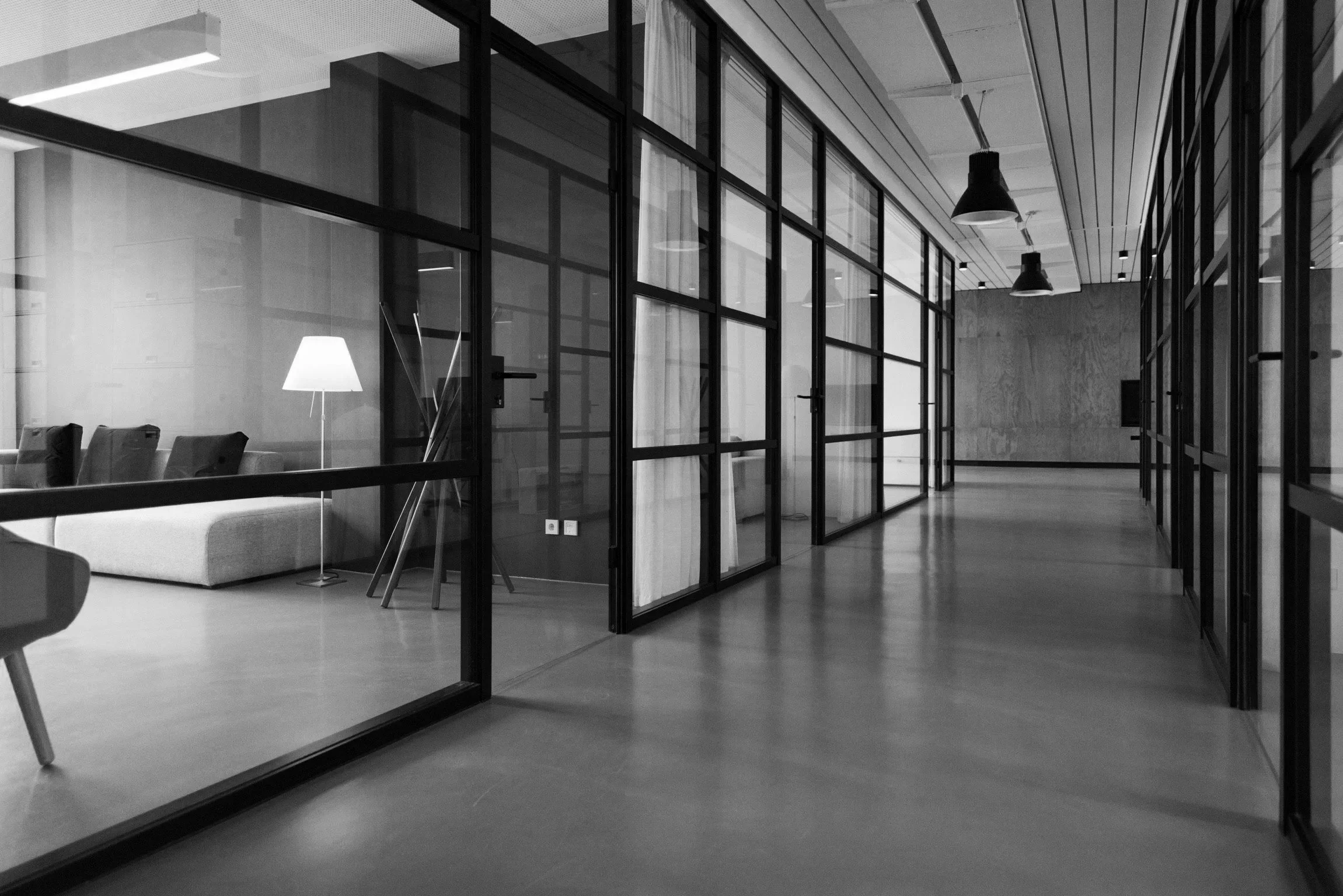 Modern office corridor with glass partitions and wooden flooring, furniture, and ceiling lights.