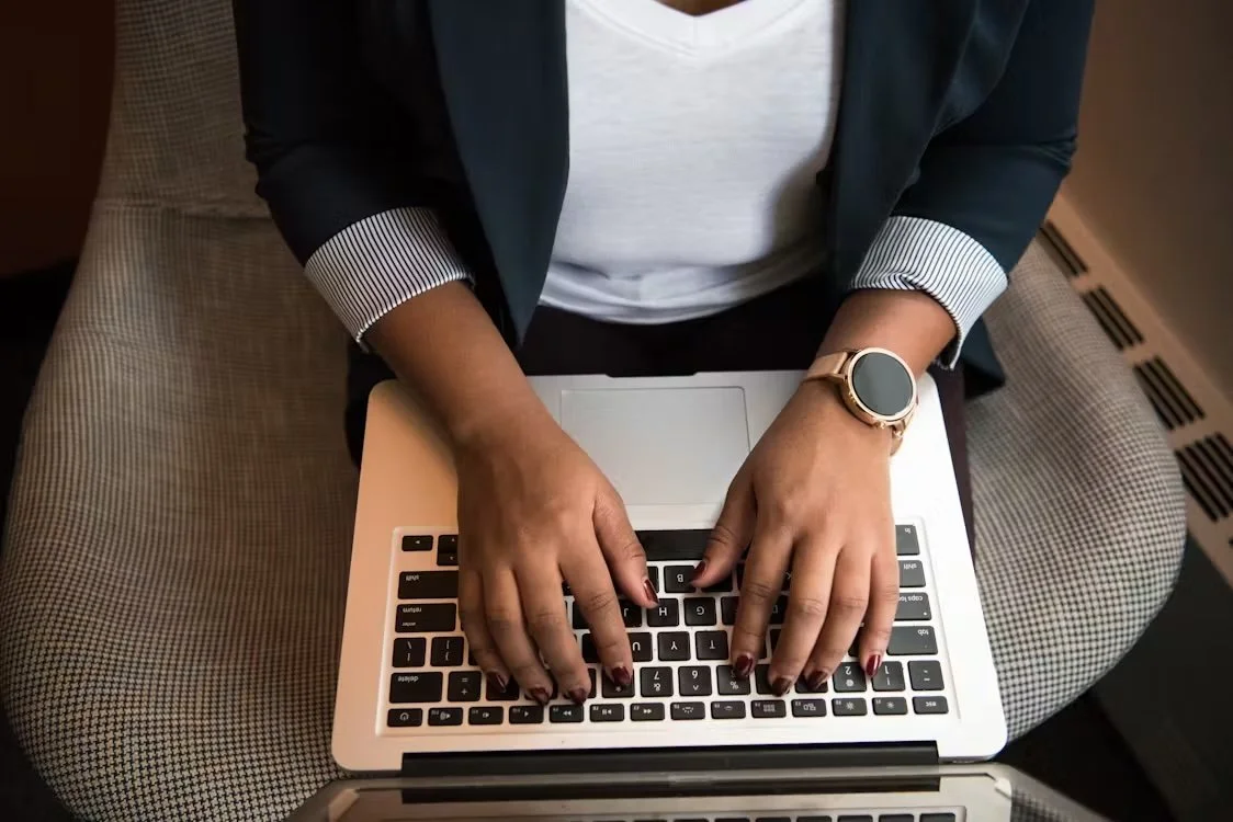 Person sitting at a table using a laptop, wearing a smartwatch and a blazer, with patterned pants.