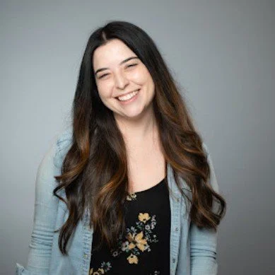 Smiling woman with long dark hair, wearing a floral top and light blue cardigan, against a neutral gray background.