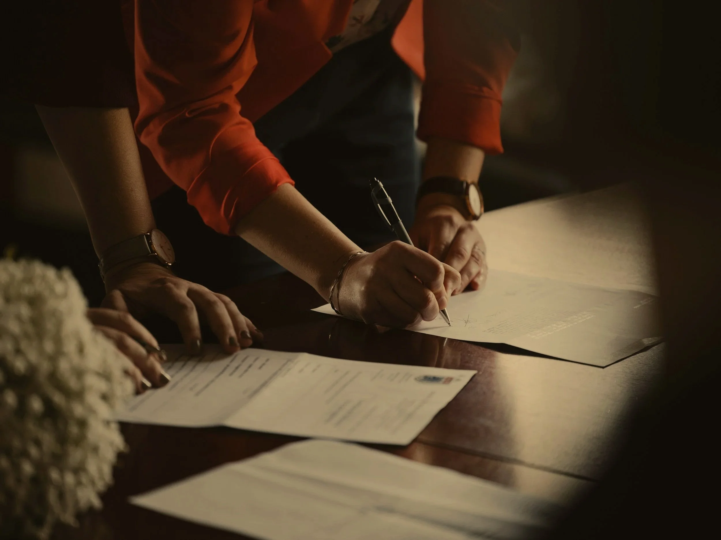Person signing a document on a wooden table with other documents nearby, in a dimly lit environment.
