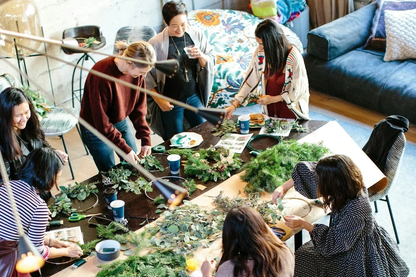 Group of women making Christmas wreaths around a large table decorated with greenery, ribbon, and decorations in a cozy living room.
