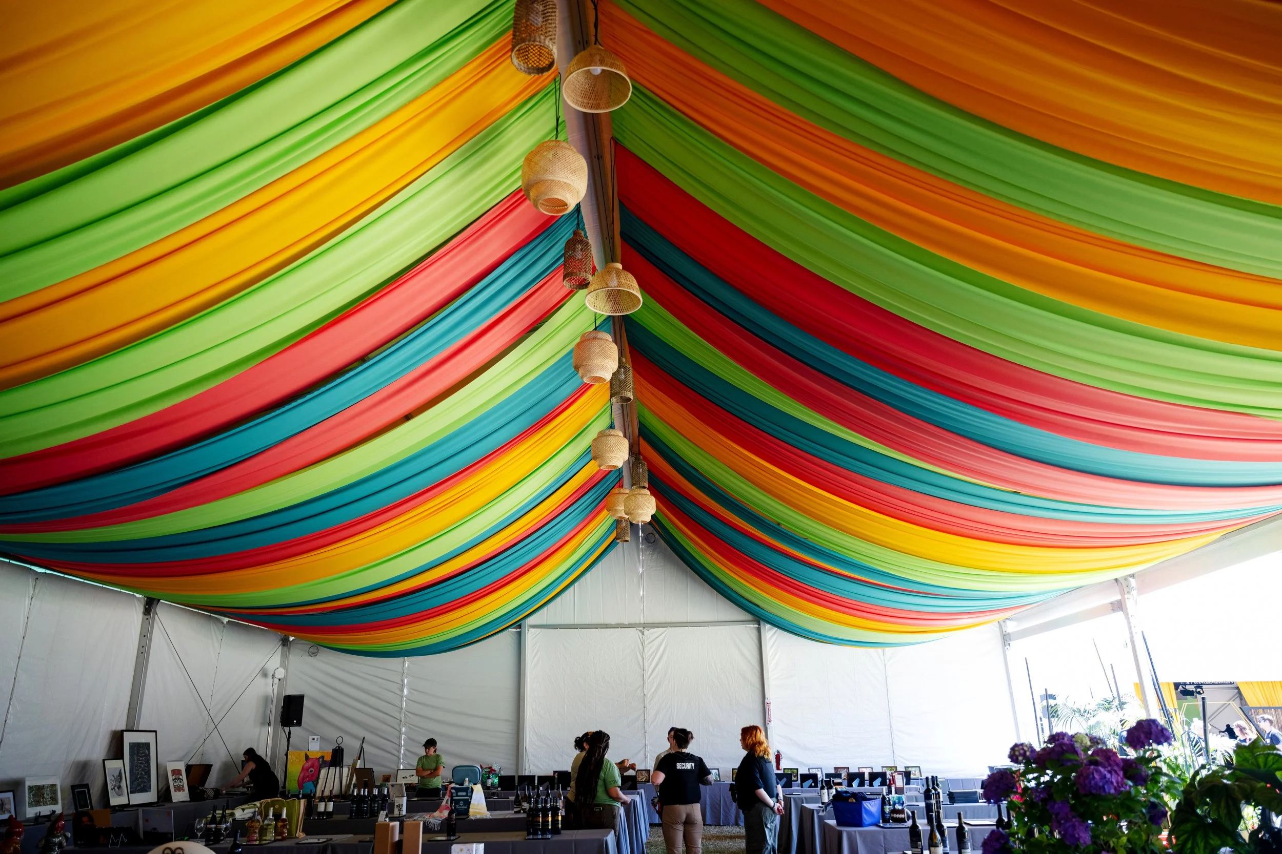 Brightly colored fabric draped across the ceiling of a large event tent, with pendant lights hanging down. Inside, there are tables with artwork and people browsing.