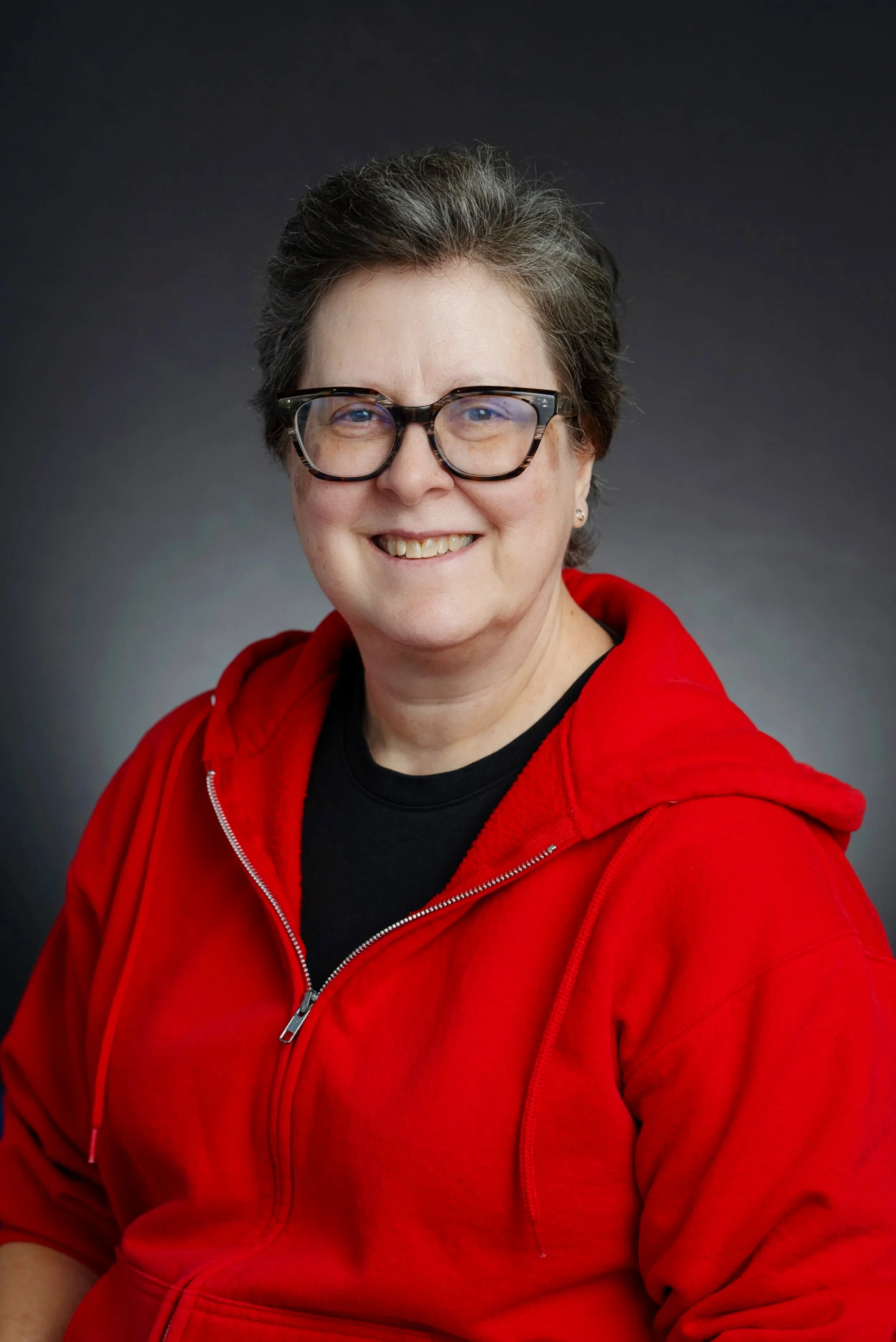 A woman with short, dark hair and glasses, smiling, wearing a black shirt and a red zip-up hoodie, against a dark gradient background.