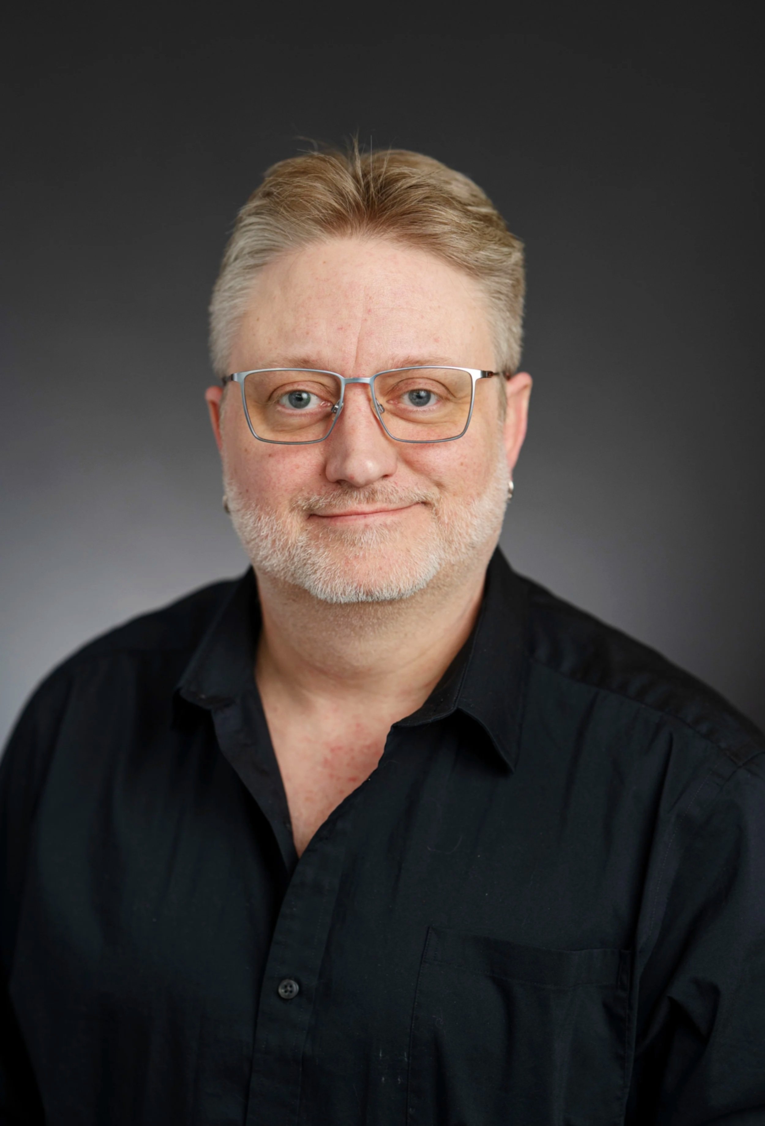 Headshot of a middle-aged man with light skin, short gray hair, blue eyes, glasses, and a light beard, wearing a black shirt and smiling lightly.