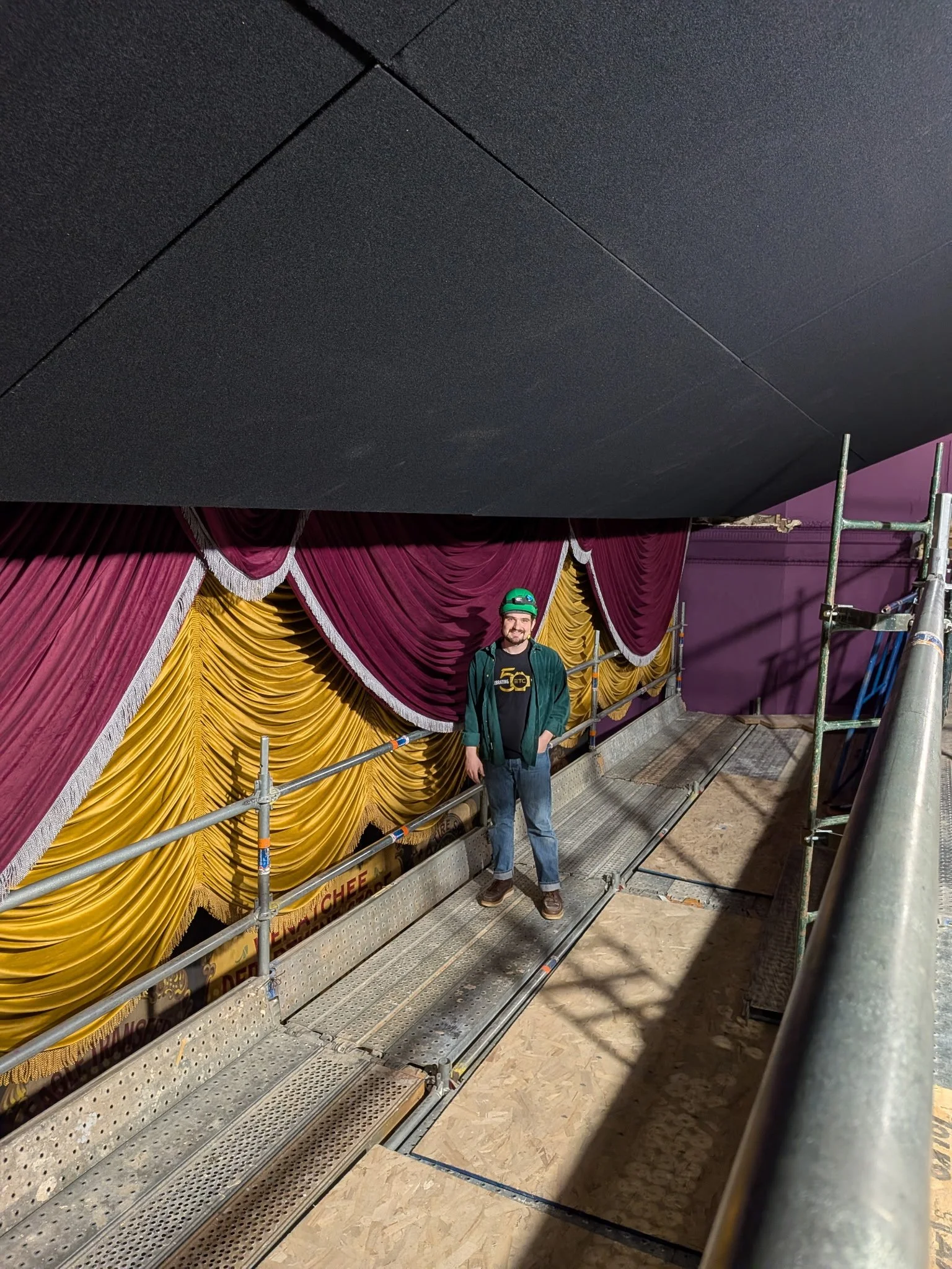 A man wearing a green helmet and green jacket stands on a metal platform in front of a stage with ornate, velvety red and gold drapes. There is scaffolding on the right side of the stage.