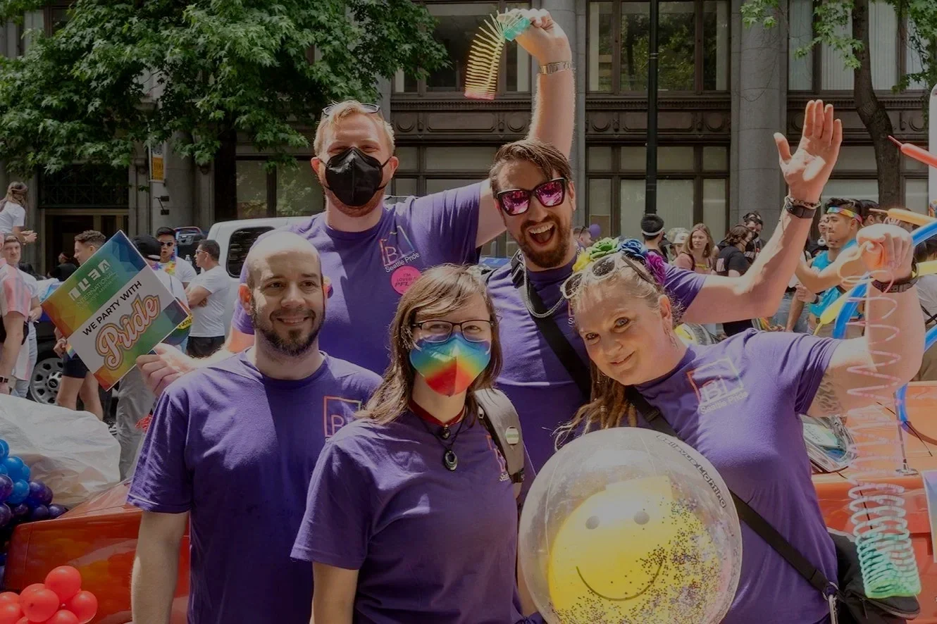 Group of five people wearing purple shirts celebrating Pride, some wearing rainbow masks, with rainbow-themed accessories and a clear balloon with a smiley face while standing near a decorated float at a Pride parade.