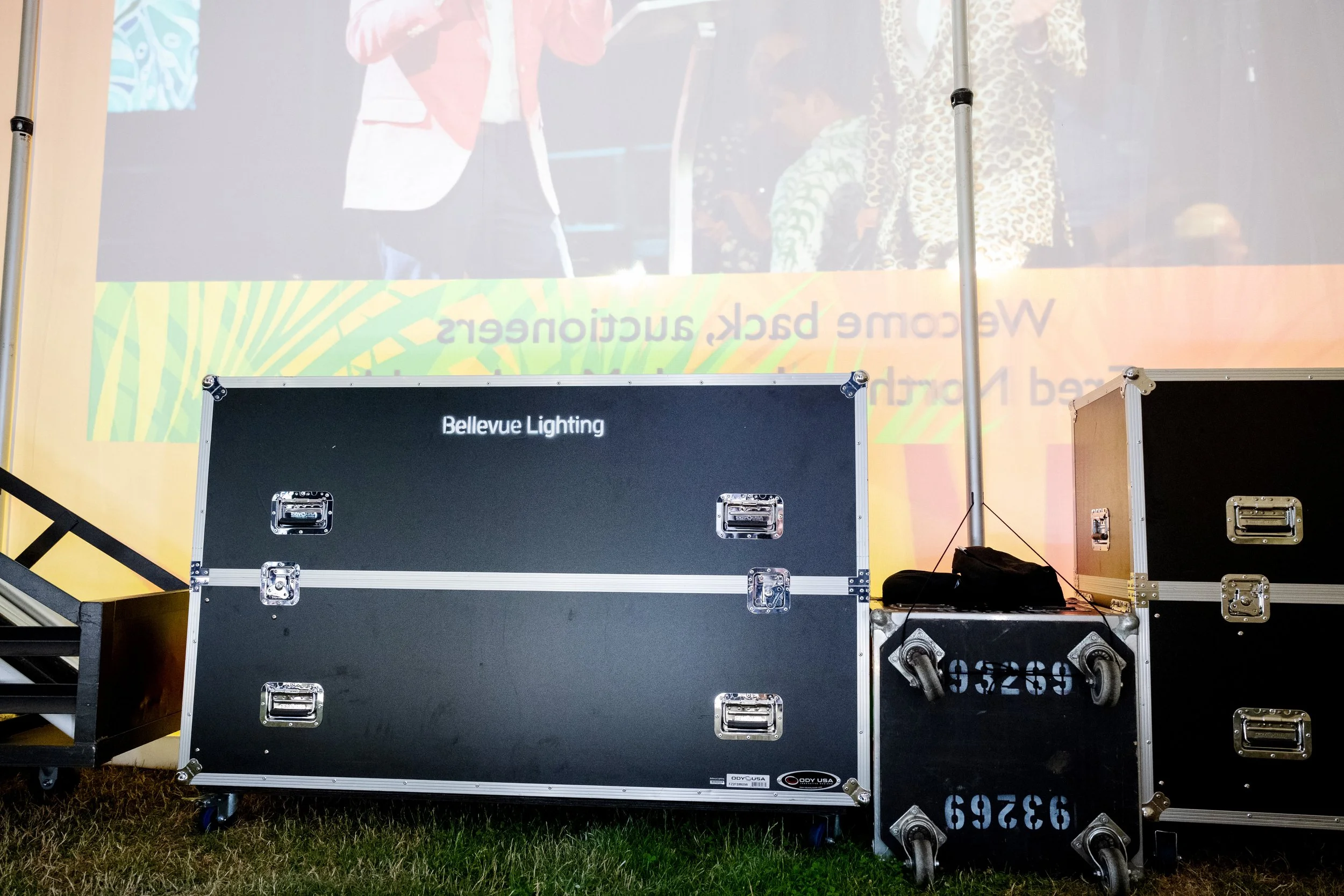 Black flight case with silver hardware and wheels, labeled 'Bellevue Lighting,' placed on a grassy area in front of a large screen or banner, with smaller cases stacked nearby.