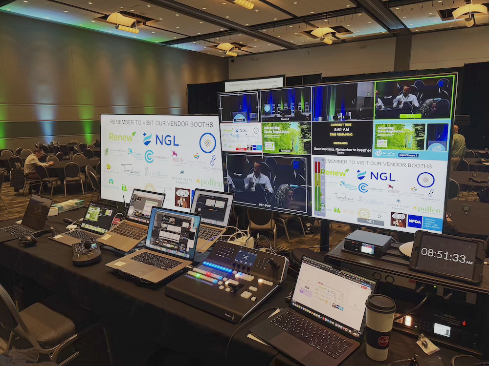 Conference room with multiple laptops and large screens displaying event information, vendor booth advertisements, and a clock showing 8:51 AM.