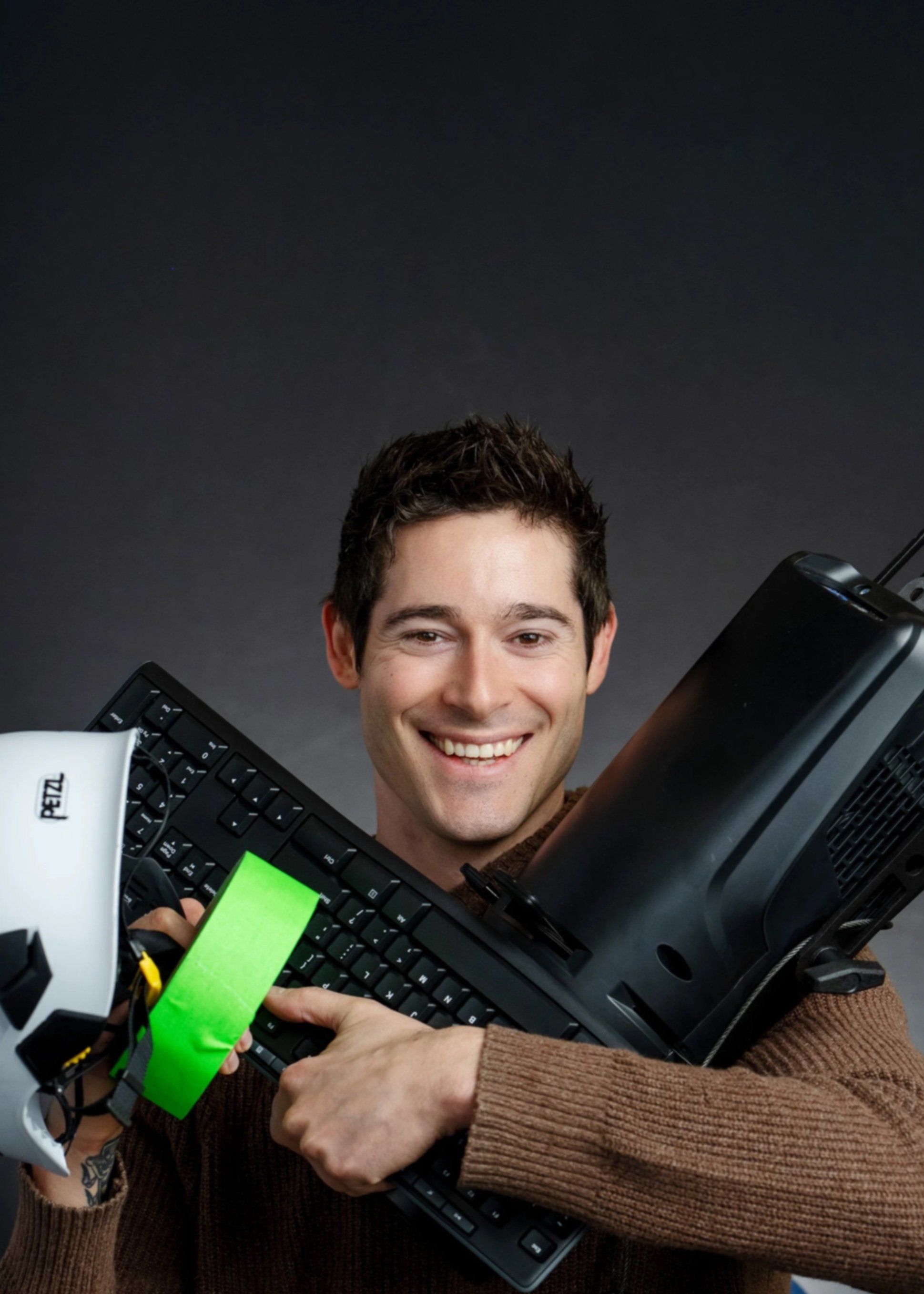 A close-up of a man with short dark hair smiling, holding multiple electronic devices including a keyboard, a gaming controller, and a laptop, all held together in one arm against a dark background.