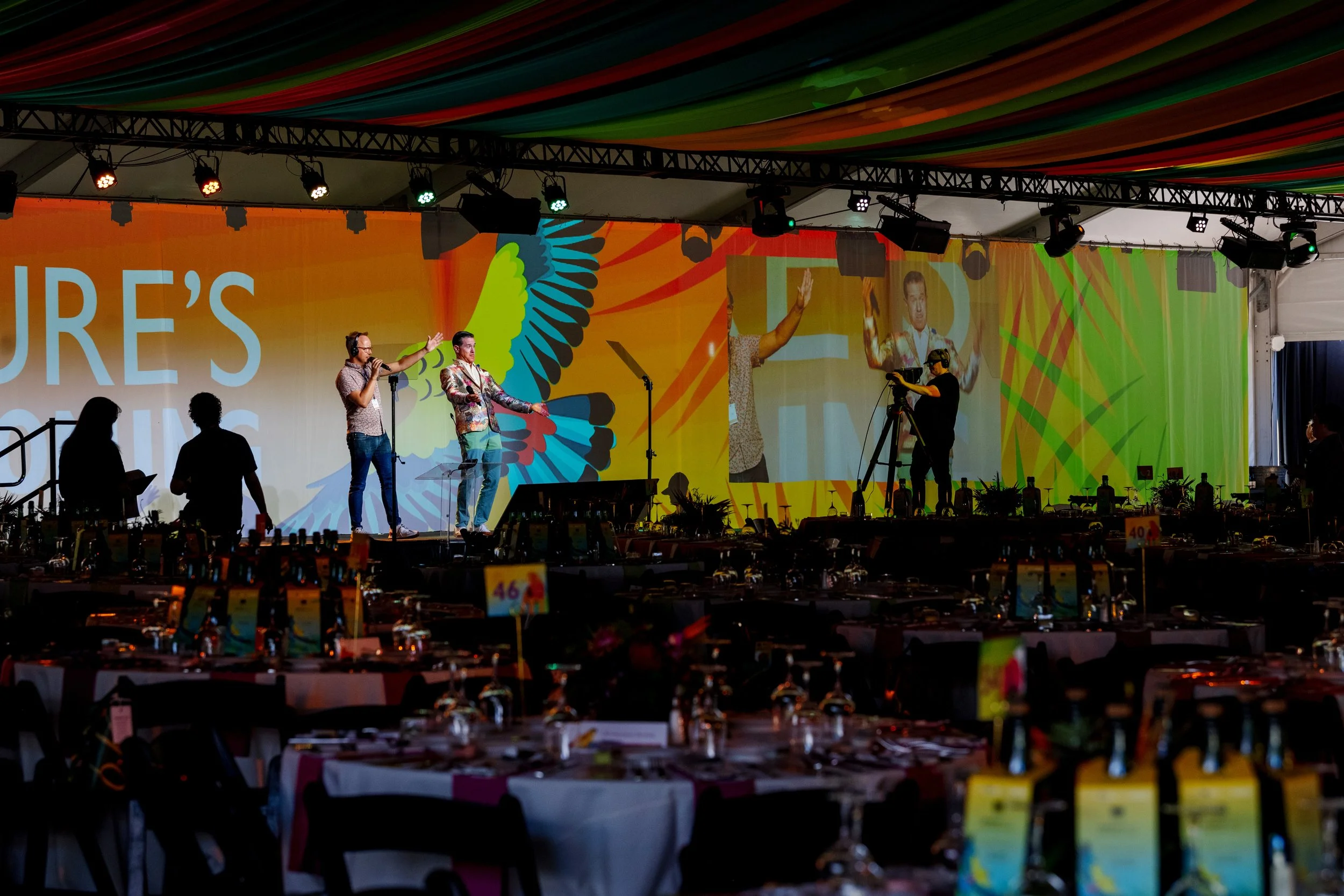 Stage with four people speaking and a cameraman filming, colorful backdrop with abstract designs, tables with merchandise or auction items in foreground.