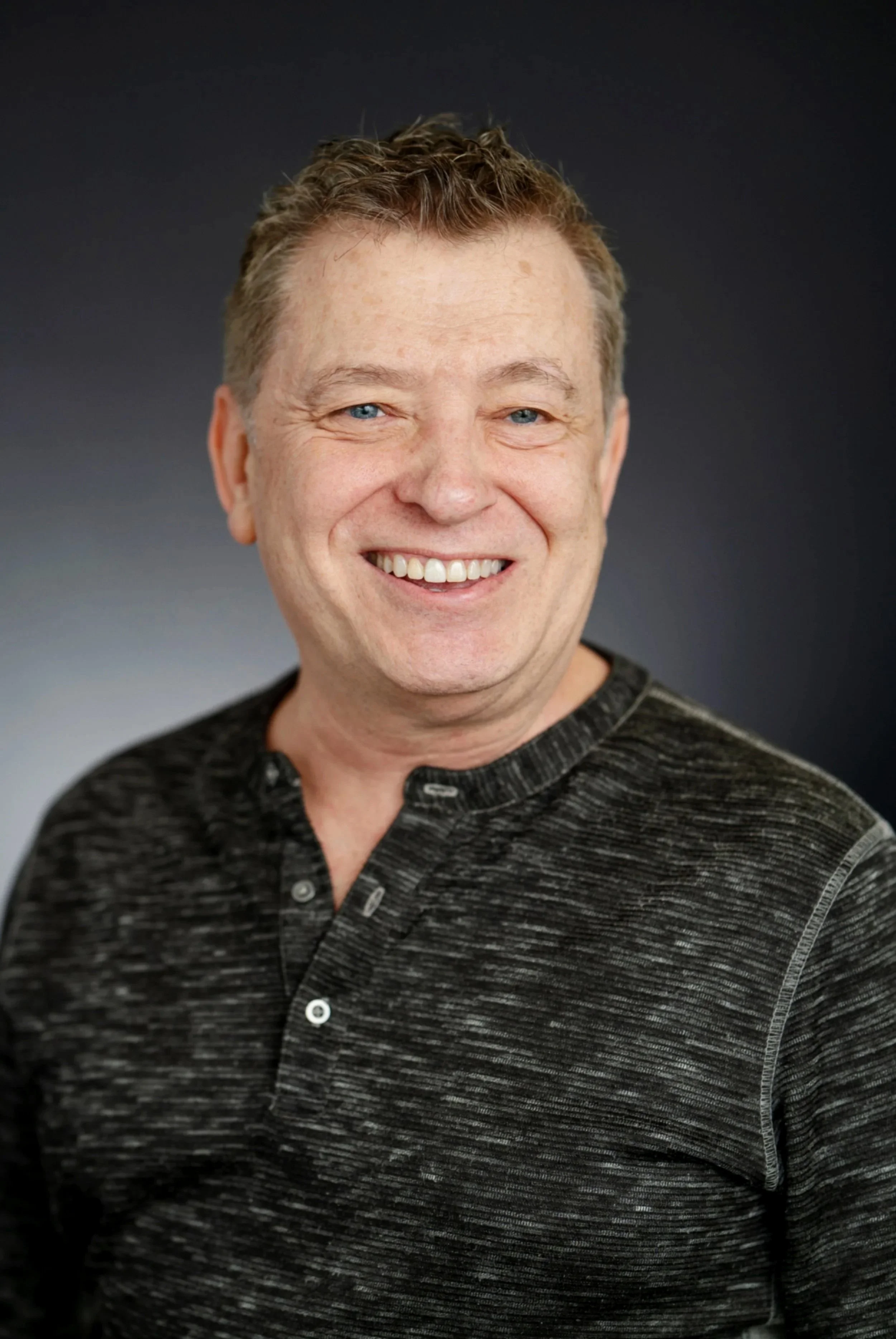 A smiling man with short curly brown hair, blue eyes, and a fair complexion, wearing a black and gray textured button-up shirt, against a dark gradient background.