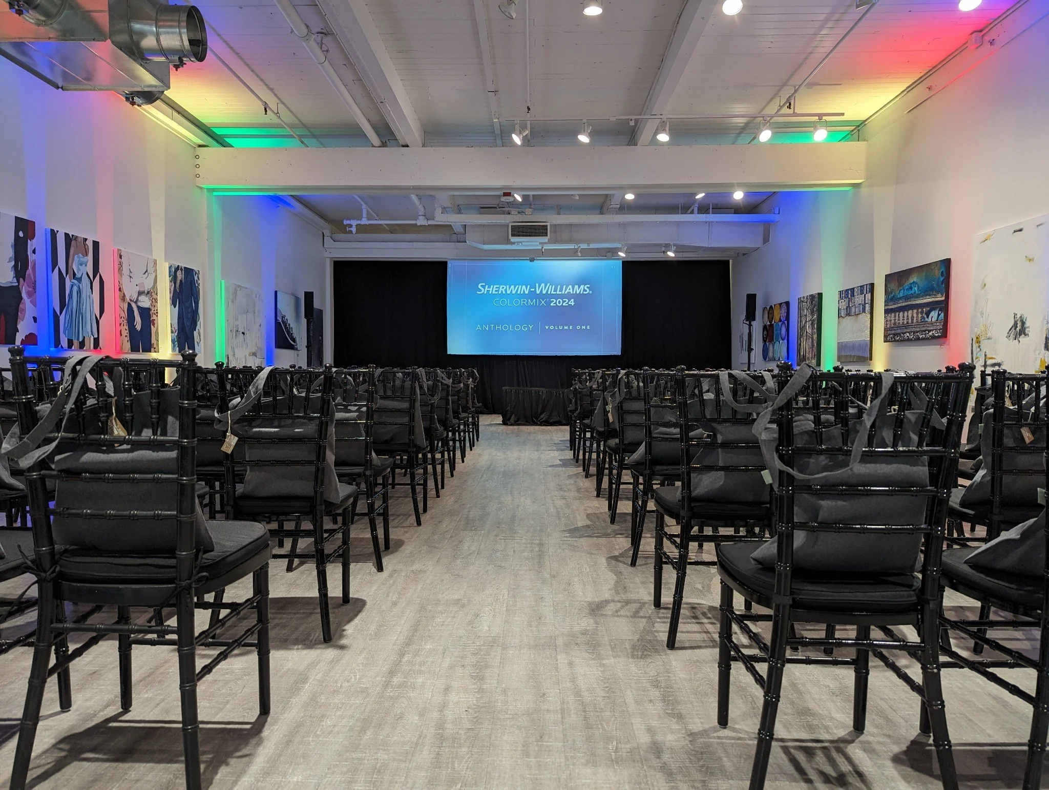 An event space or gallery with chairs arranged in rows facing a large screen that reads 'Sherwin-Williams ColorMix 2024 Anthology Volume One.' The room is decorated with colorful lighting and artwork on the walls.