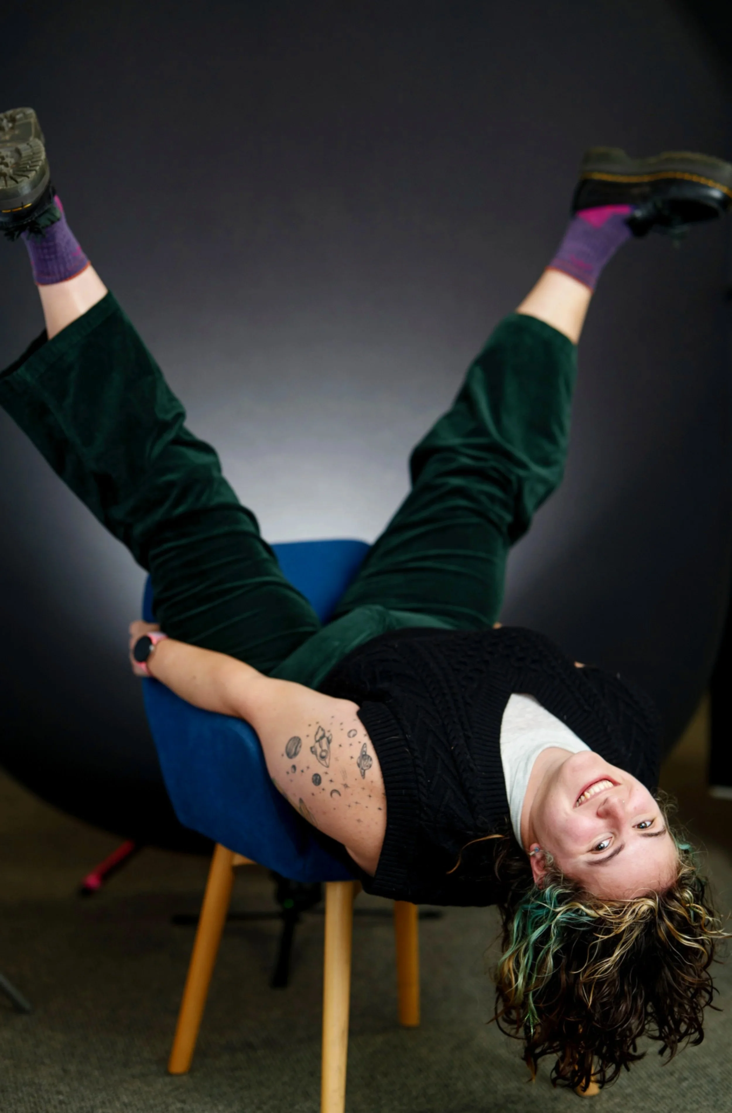 A woman with tattoos of planets and astronauts lying upside down on a blue chair with her head hanging backwards and smiling.