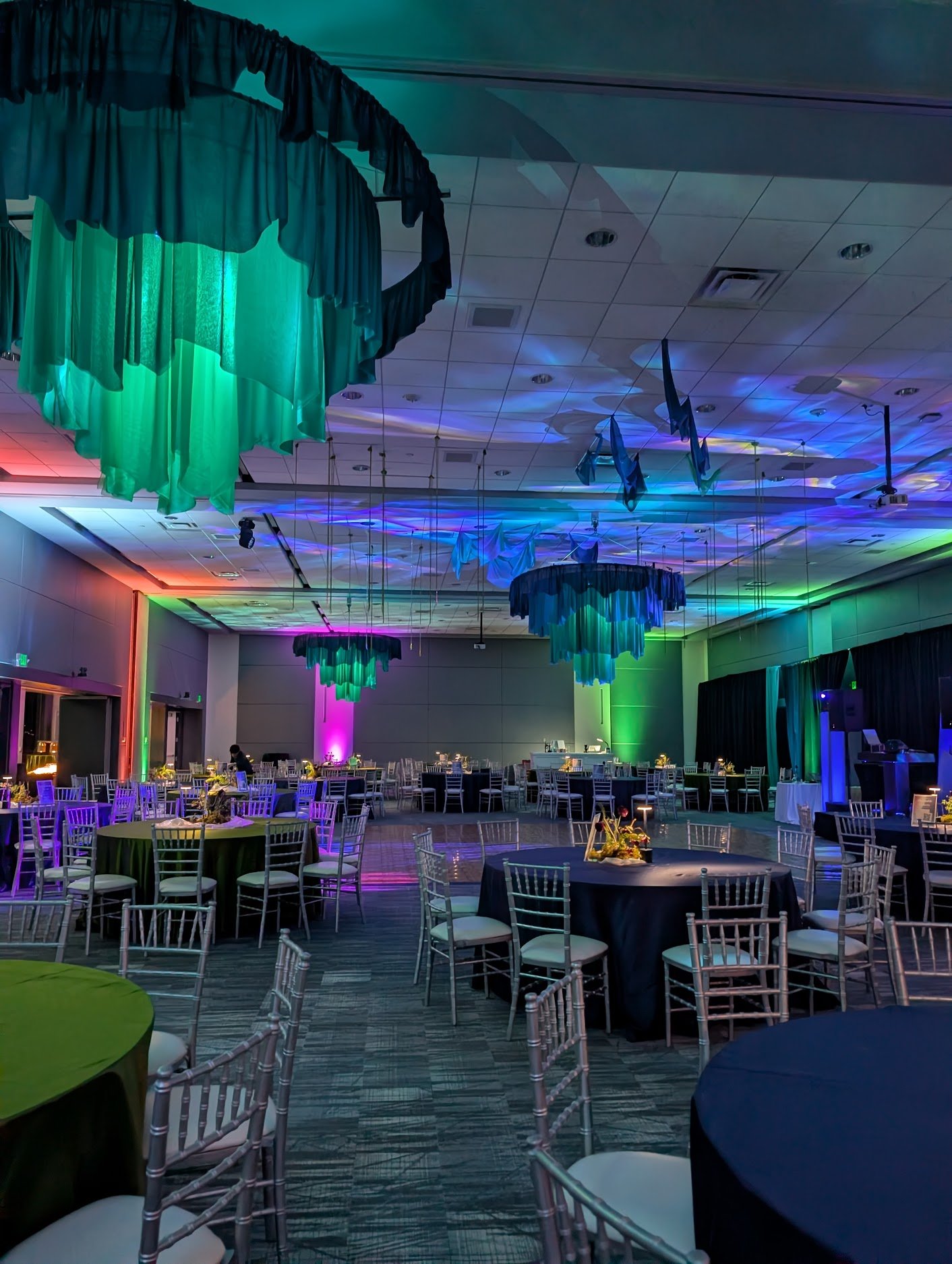 Event venue decorated with colorful lighting, draped fabric on the ceiling, round tables with black tablecloths, and white chairs arranged around the tables.