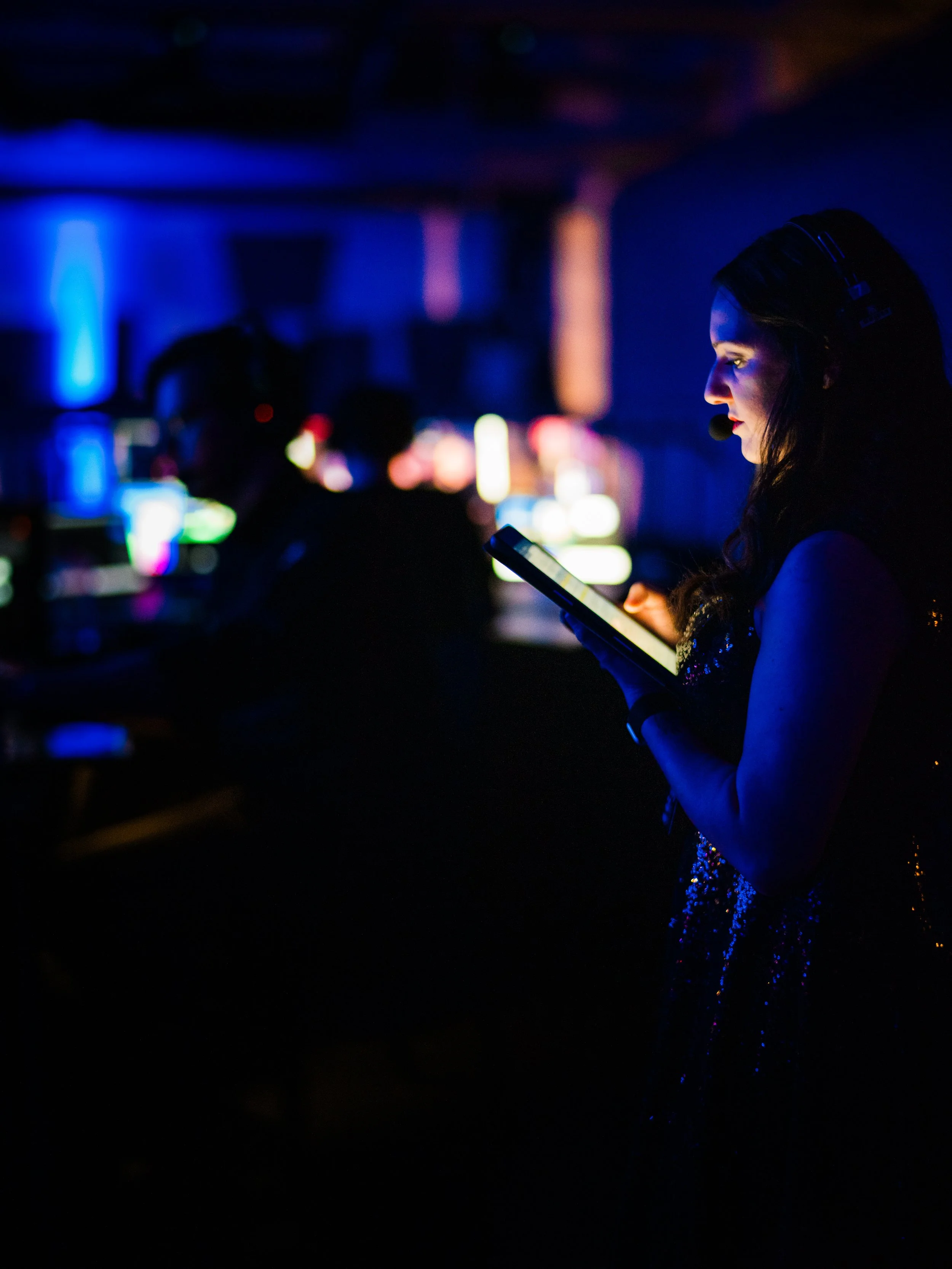 A woman with dark hair, wearing a sequined dress, is holding a tablet and wearing a headset with a microphone, illuminated by blue and purple stage lights, with blurred figures and colorful lights in the background.