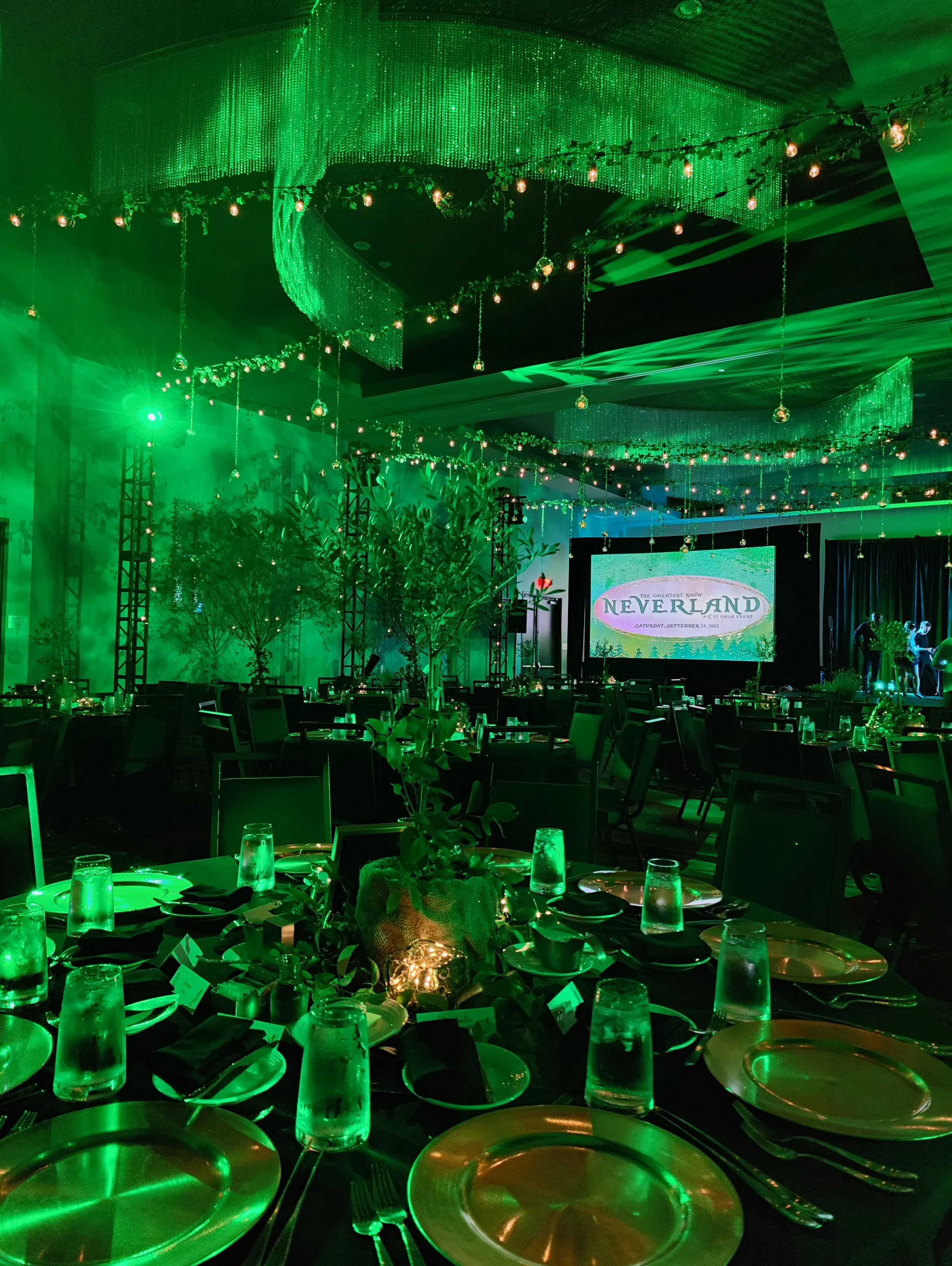 Indoor event space decorated with green lighting, hanging string lights and decorations, tables set with plates, glasses, and utensils, and a large screen displaying the logo for the Neverland event.