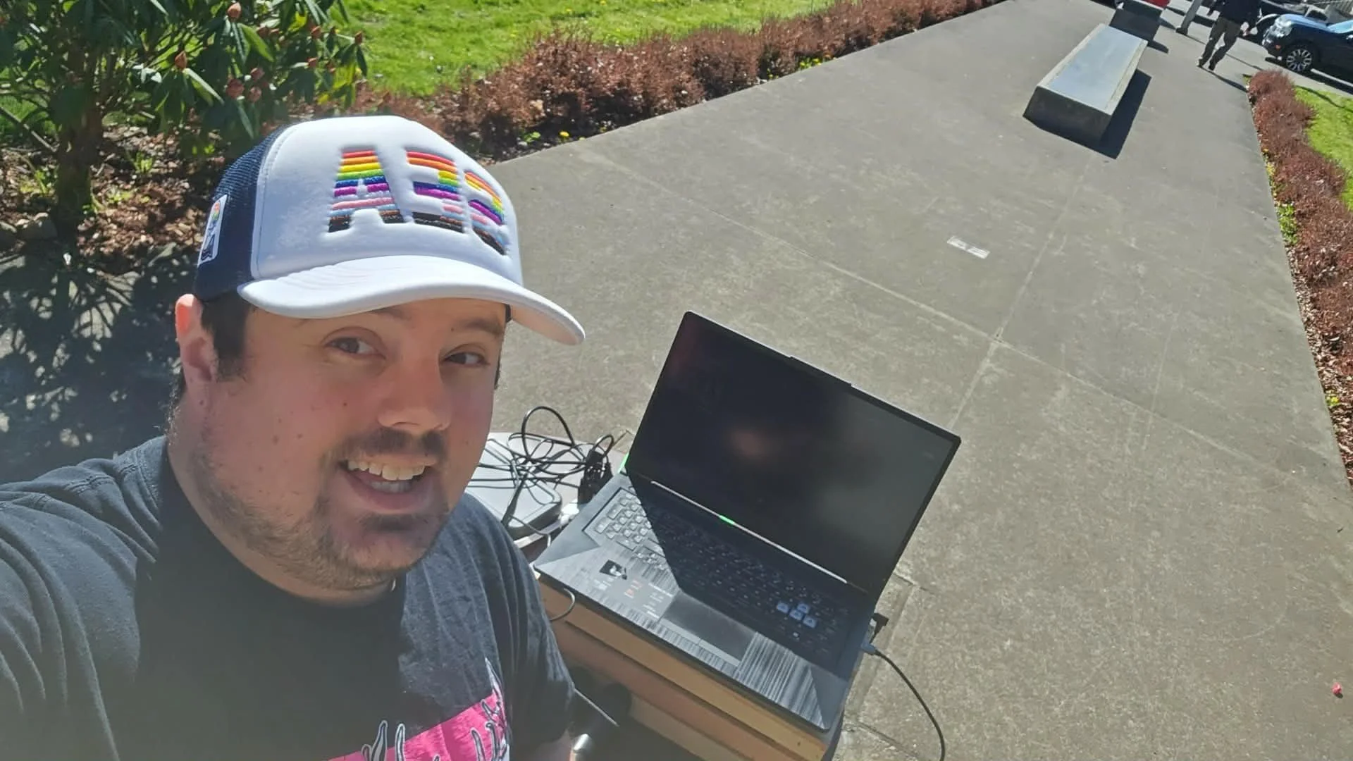 A man takes a selfie outdoors on a sunny day. He is wearing a baseball cap with rainbow text and has a laptop on a cardboard box in front of him. He appears happy as he shows his teeth and is positioned near a sidewalk, with a tripod and some wires nearby.