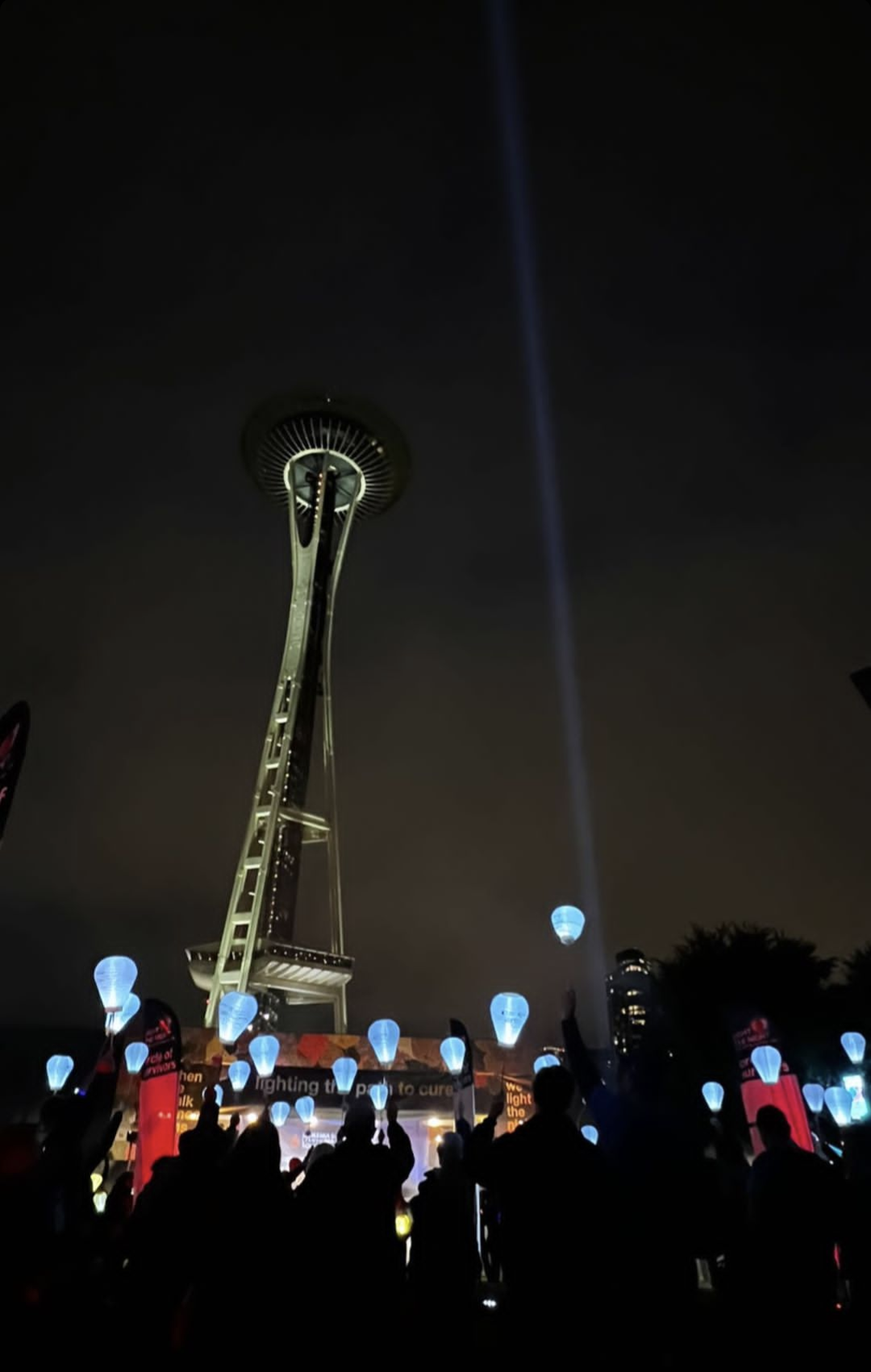 Nighttime scene in front of the Space Needle in Seattle with a crowd holding blue lanterns and a blue spotlight beam in the sky.
