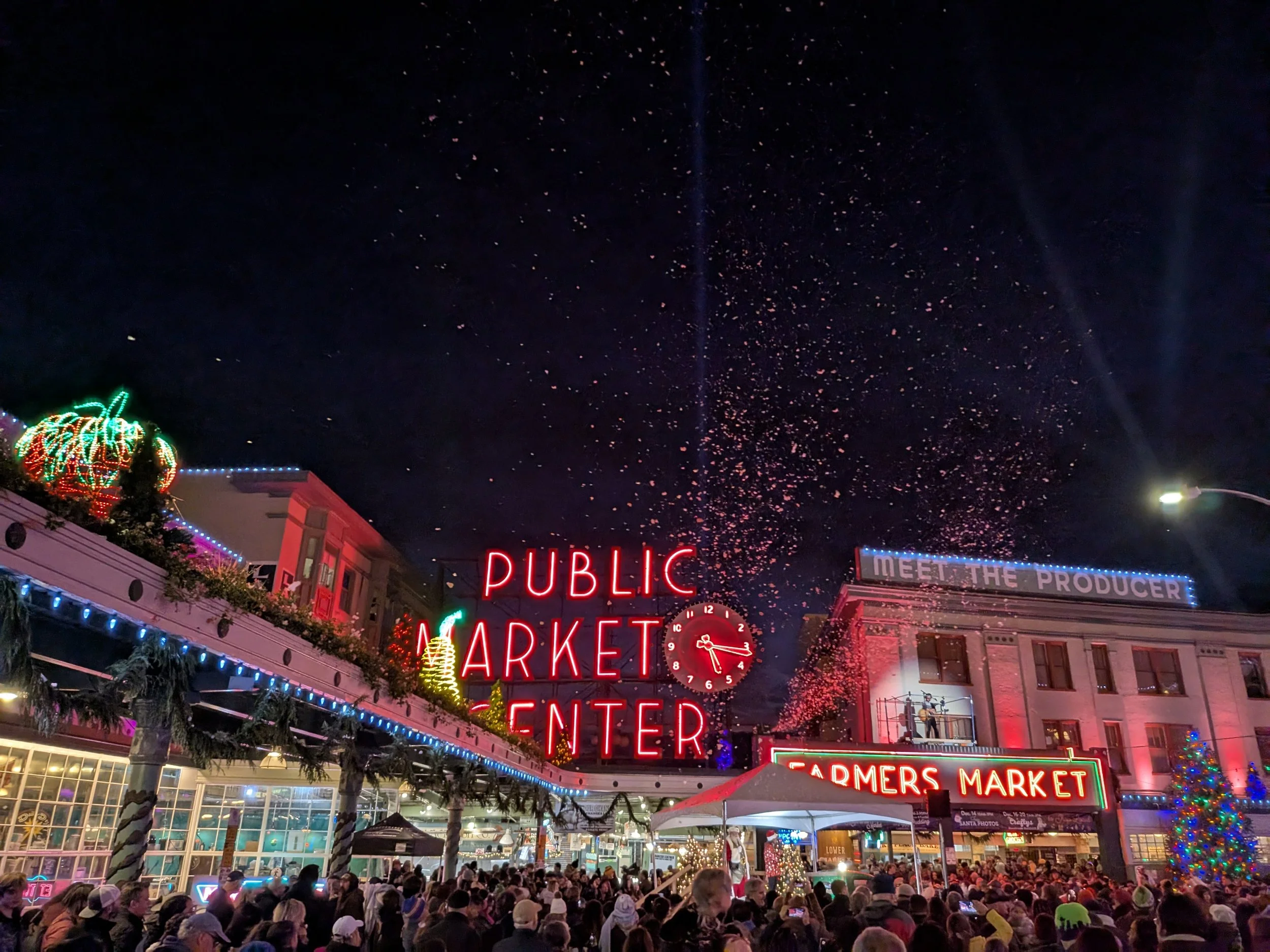 Night scene of a busy outdoor market with colorful neon and string lights, crowded with people. The storefronts display signs reading 'PUBLIC MARKET' and 'FARMERS MARKET', with a gazebo decorated with holiday lights and a large crowd gathered underneath.