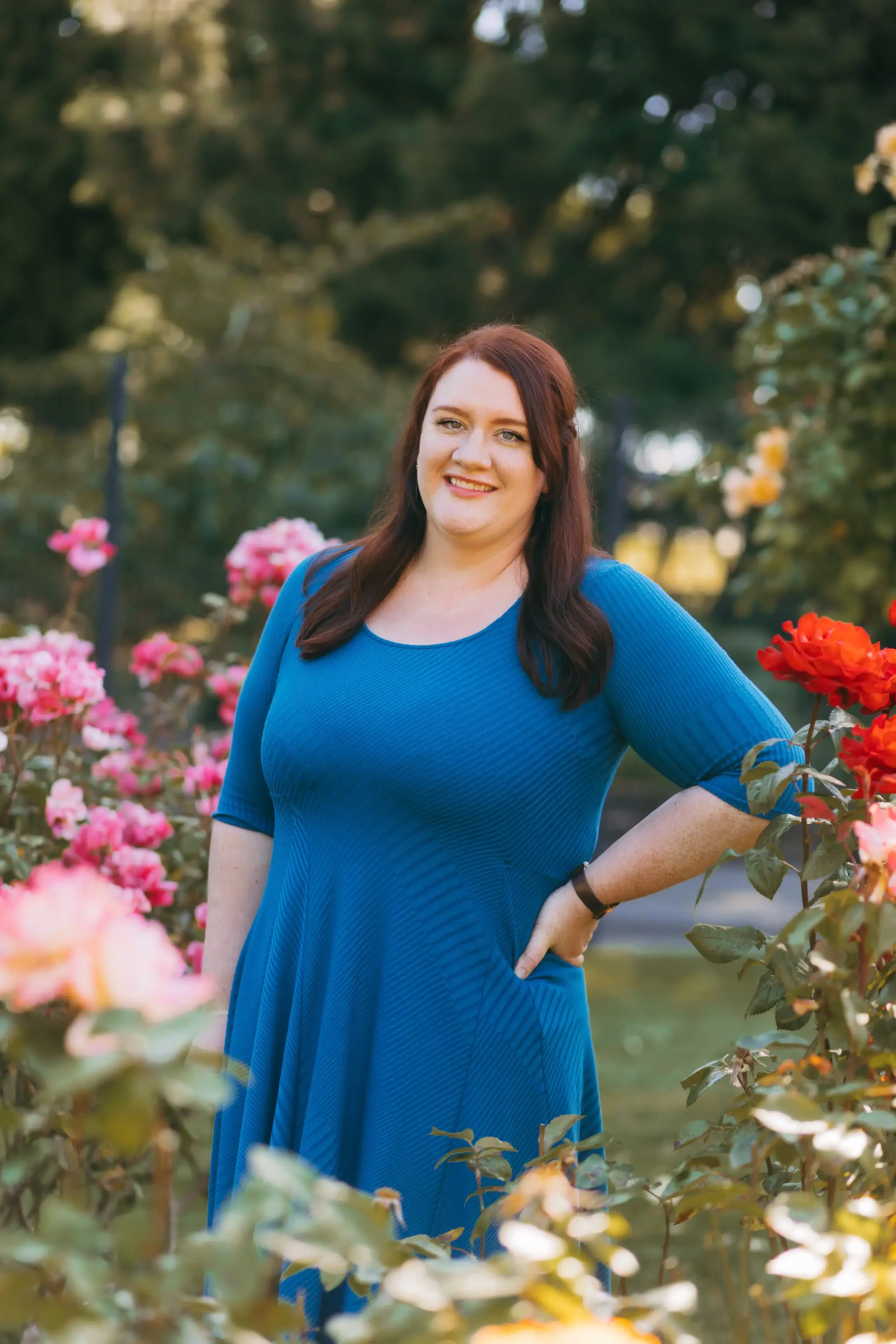 A woman with red hair, wearing a blue dress, smiling while standing in a garden with pink and red flowers, and green trees in the background.