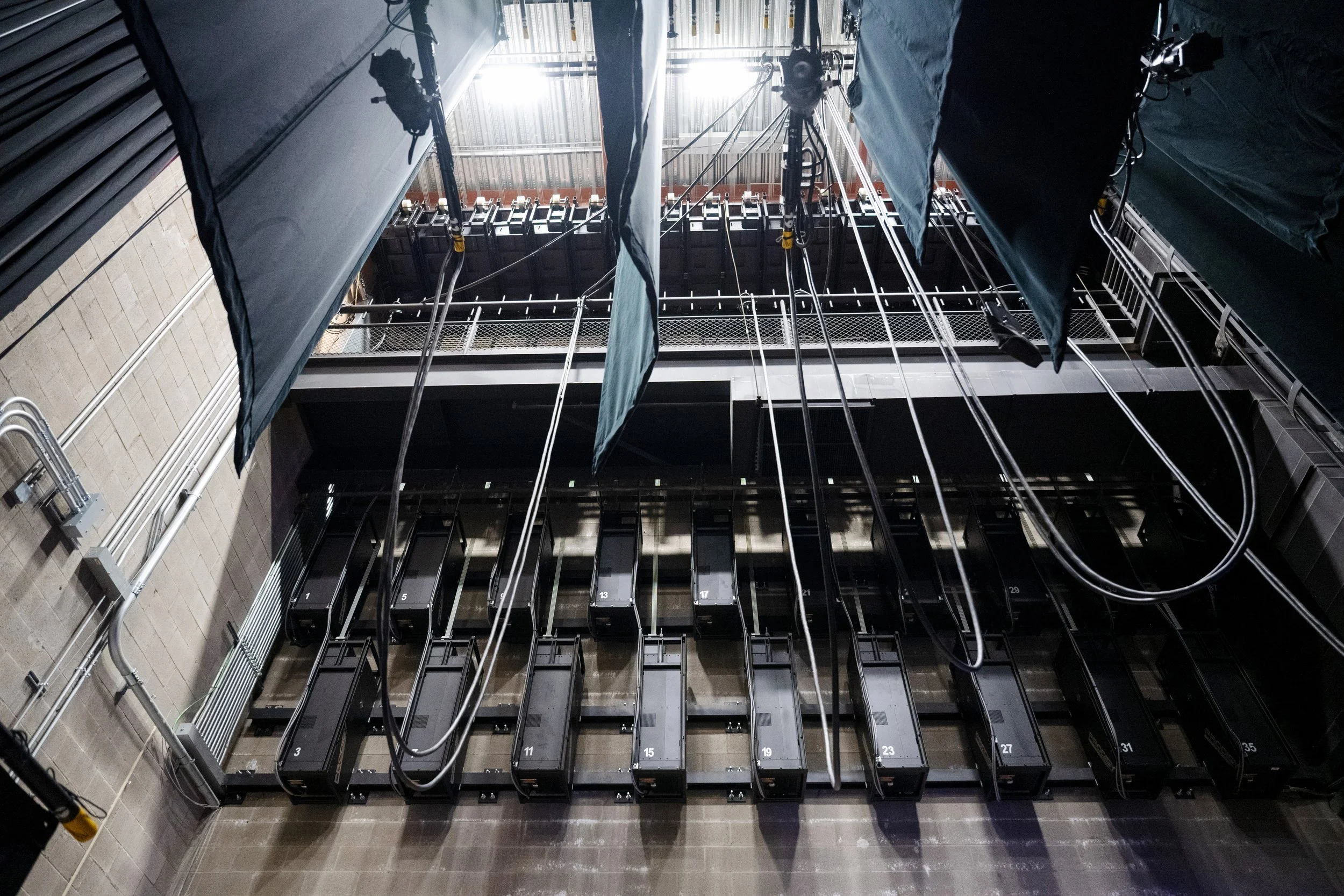 An elevator shaft with empty elevator cars numbered 1 to 35, viewed from below, with black curtains hanging and machinery above, in an industrial setting.