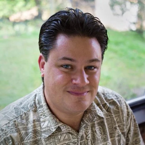 A man with dark, slicked-back hair and light skin, wearing a patterned shirt, smiling outdoors with greenery in the background.