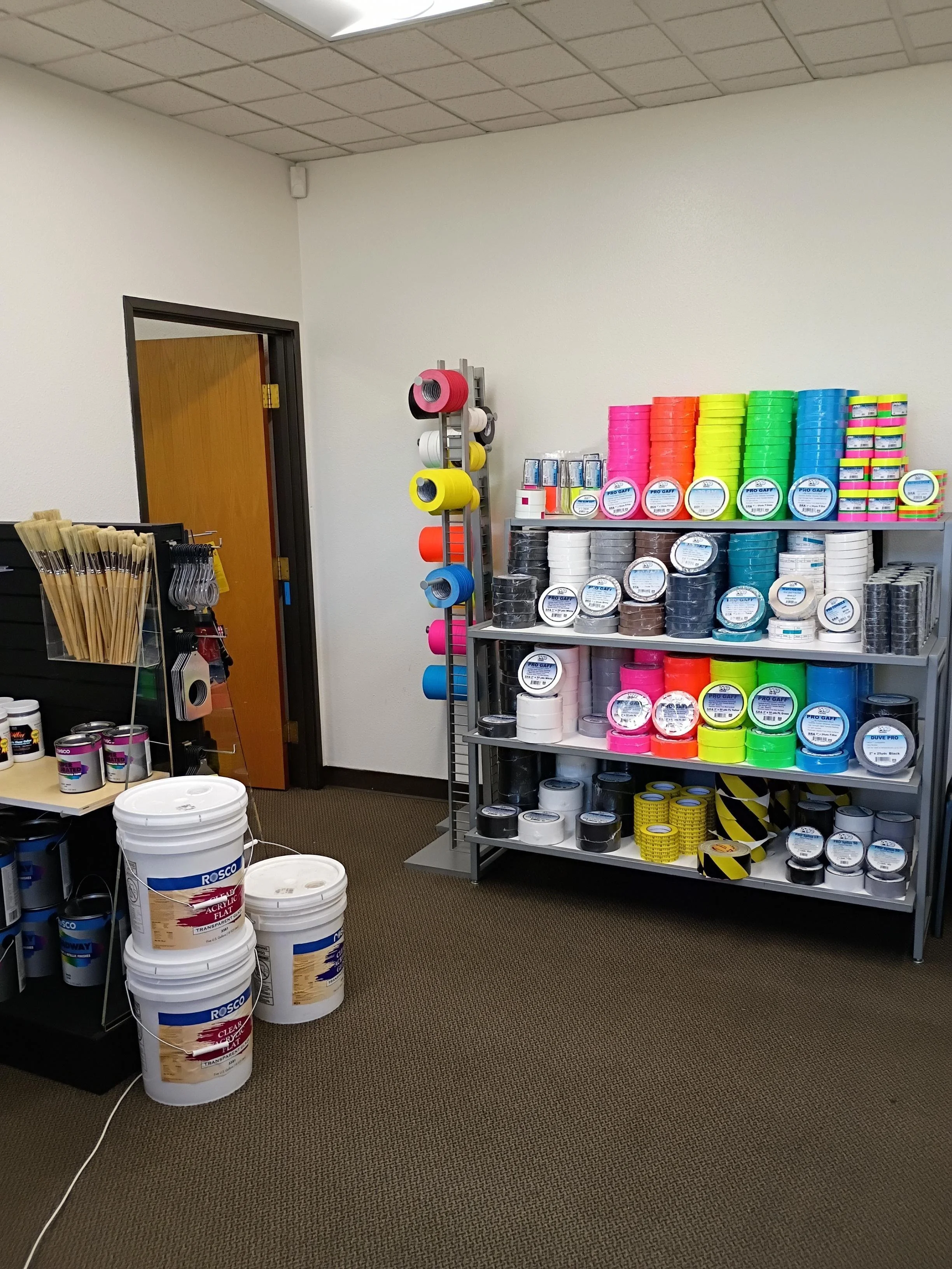 Store display with rolls of tape in various colors on shelves, buckets of paint on the floor, and brushes and tools on a table.