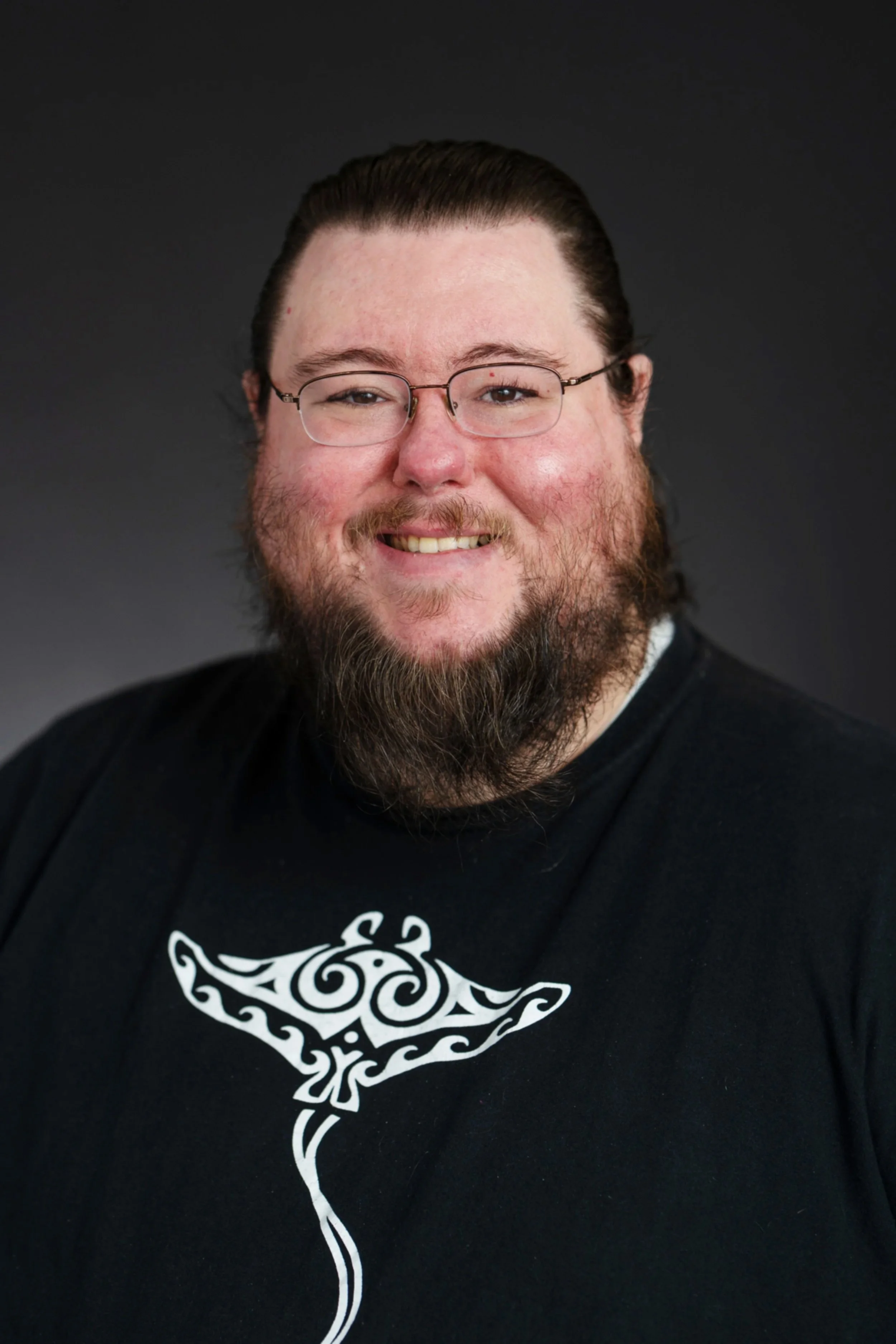 A man with glasses, a beard, and long hair tied back, smiling against a dark background, wearing a black shirt with a white tribal-style design.
