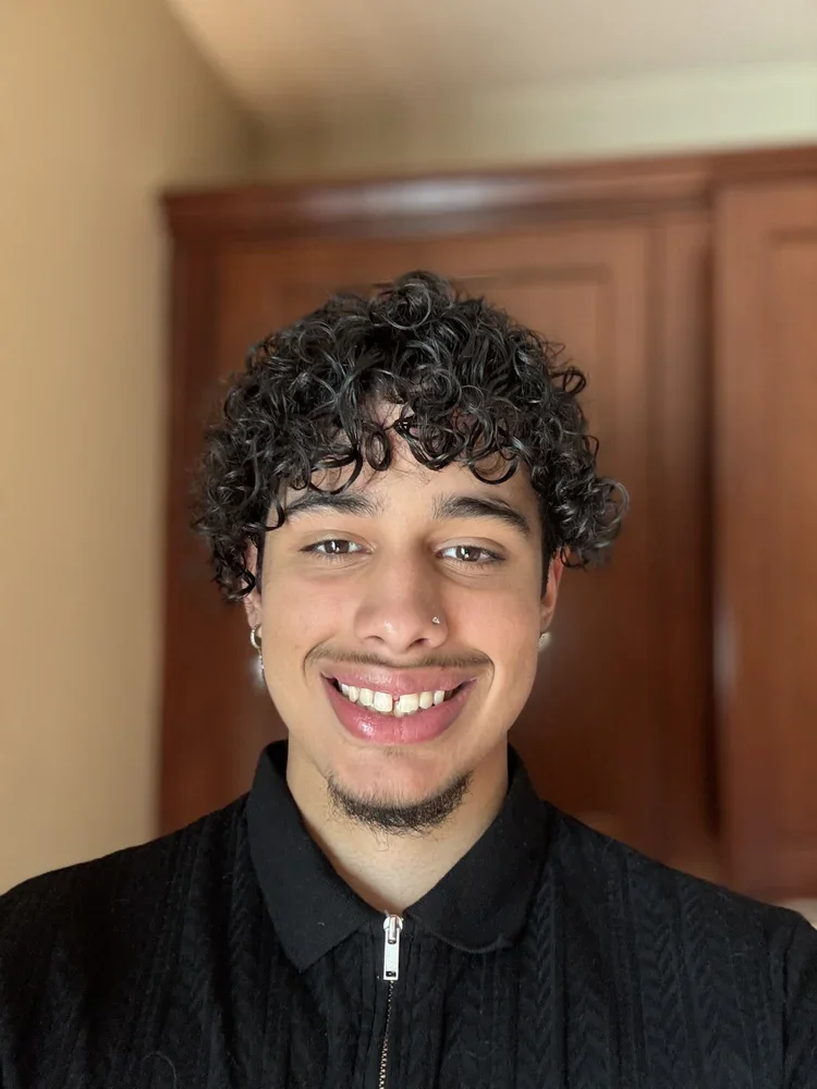 A young person with curly dark hair, light skin, a nose piercing, earrings, a goatee, and a bright smile, wearing a black zip-up jacket in front of a wooden closet.