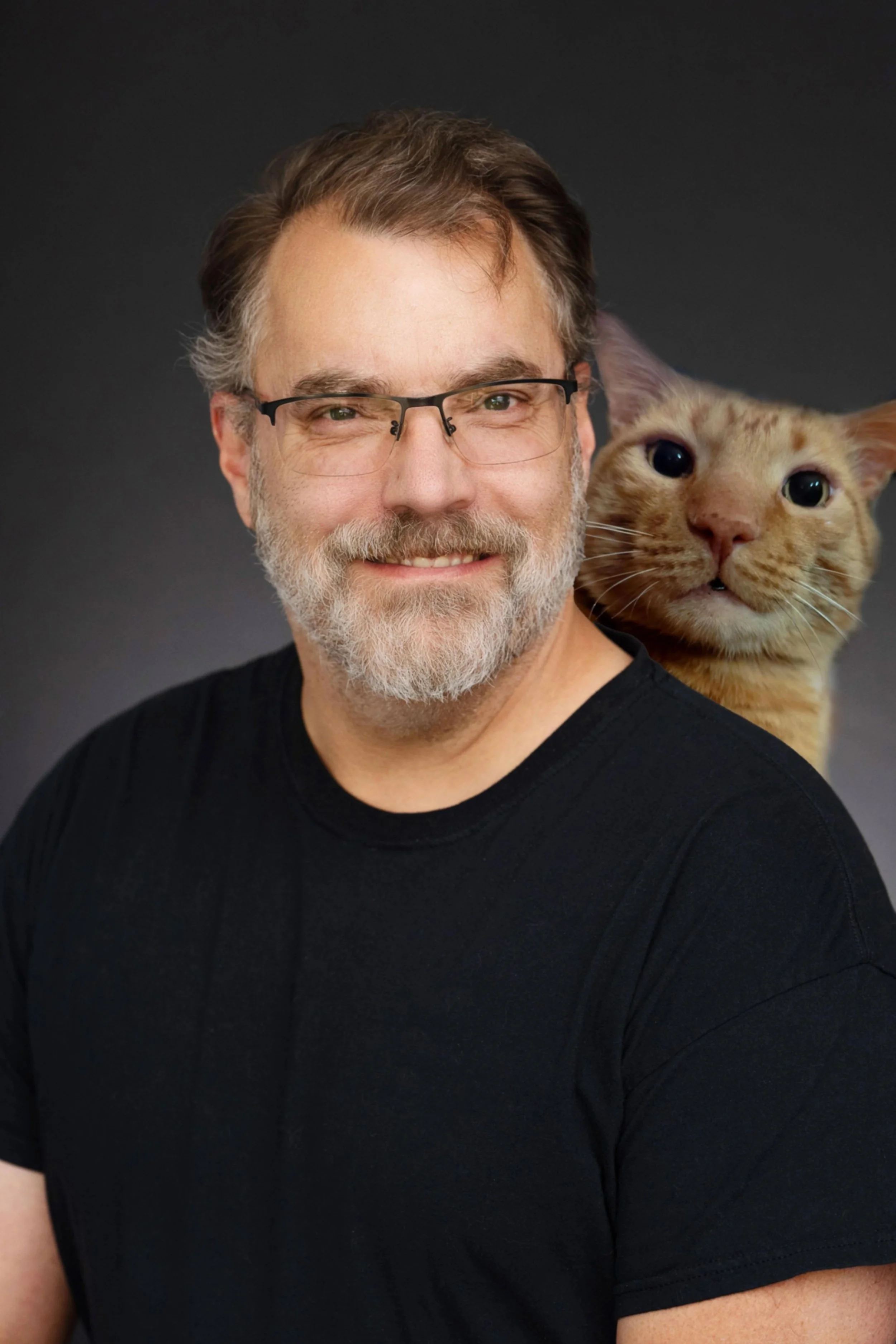 A middle-aged man with gray hair and a beard, wearing glasses and a black shirt. He is smiling and has a cat on his shoulder, which is looking directly at the camera. The background is dark gray.
