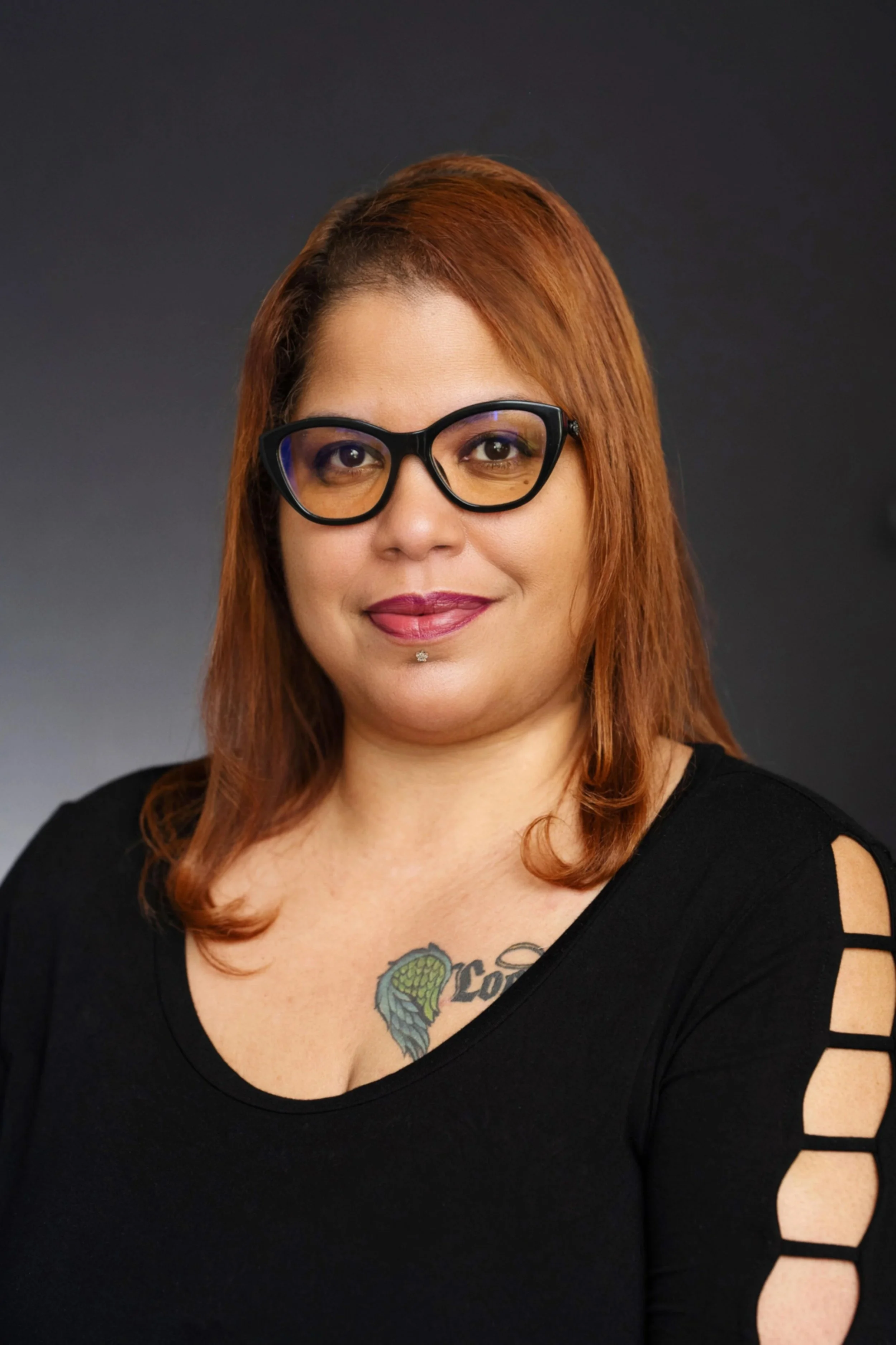 A woman with reddish-brown hair, wearing black glasses and a black top with cutout sleeves, has a tattoo of a snake emerging from a flower on her chest and is smiling against a dark grey background.