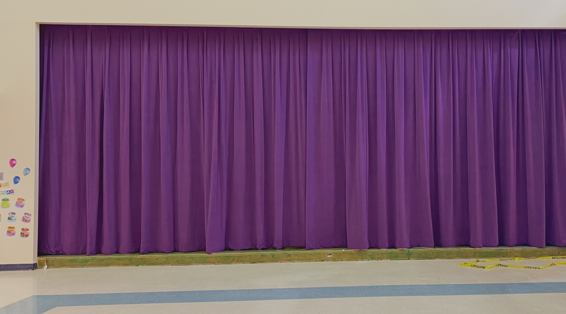 A closed purple curtain on a stage, with a partial view of a decorated wall to the left and yellow caution tape on the floor in front of the stage.
