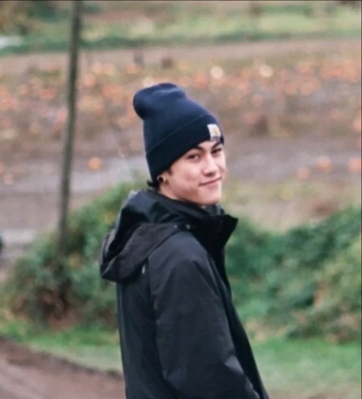 Young man wearing a navy blue beanie and black jacket, smiling outdoors on a fall day with blurred trees and grass in the background.