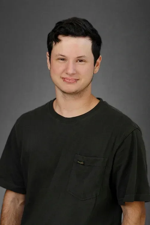 Young man with short dark hair, wearing a black t-shirt, smiling with his tongue slightly sticking out, against a gray background.