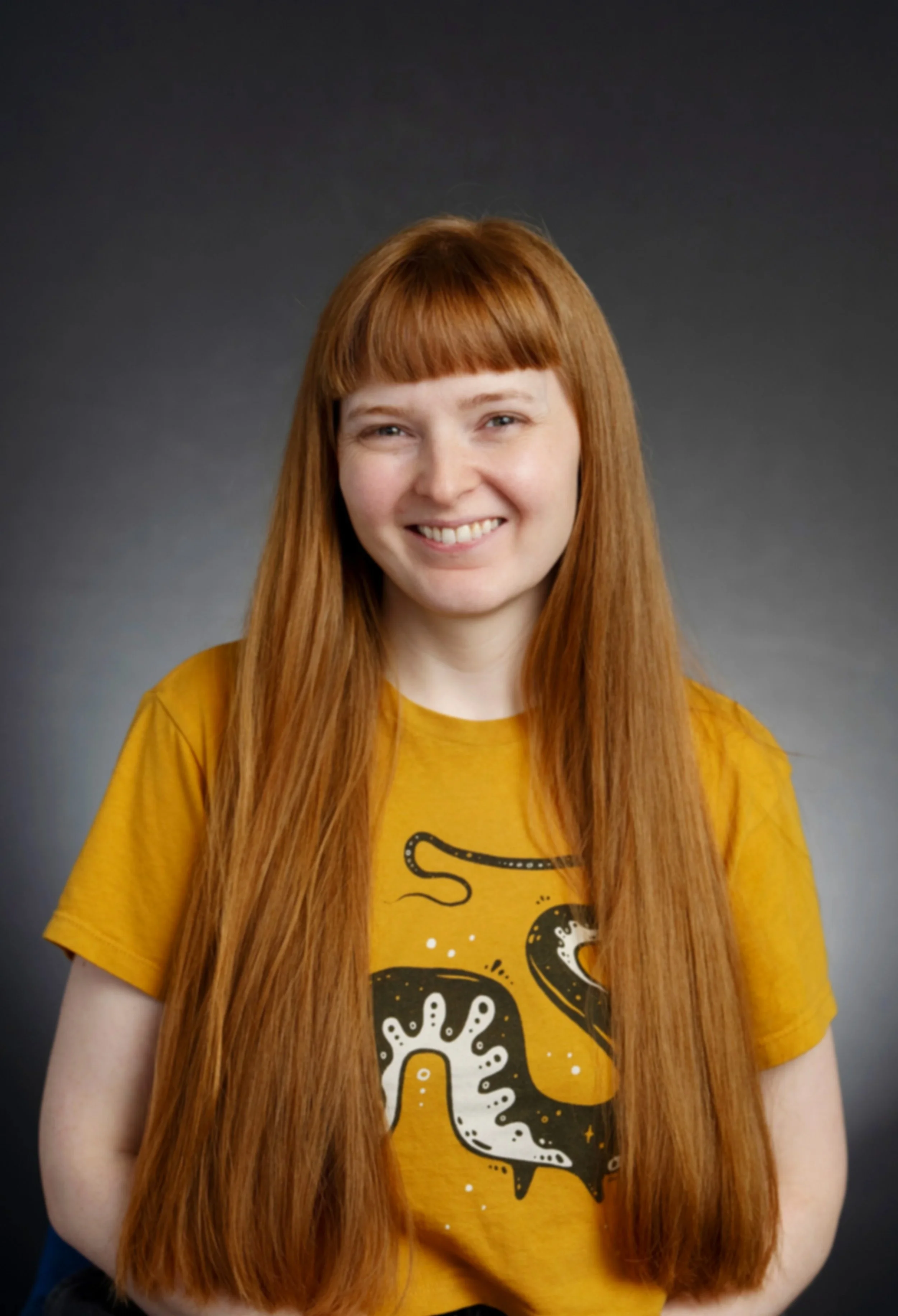 A young woman with long red hair, smiling, wearing a yellow T-shirt with cartoon octopuses and dots, against a dark, plain background.