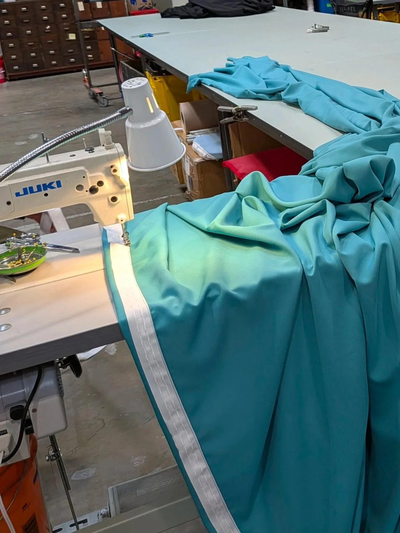 A workstation with sewing equipment, fabric, and tools in an industrial setting, including a sewing machine, blue and green fabric, a lamp, and various supplies.
