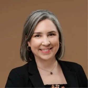 Portrait of a smiling woman with shoulder-length gray hair, wearing a black blazer, earrings, and a necklace, against a brown background.