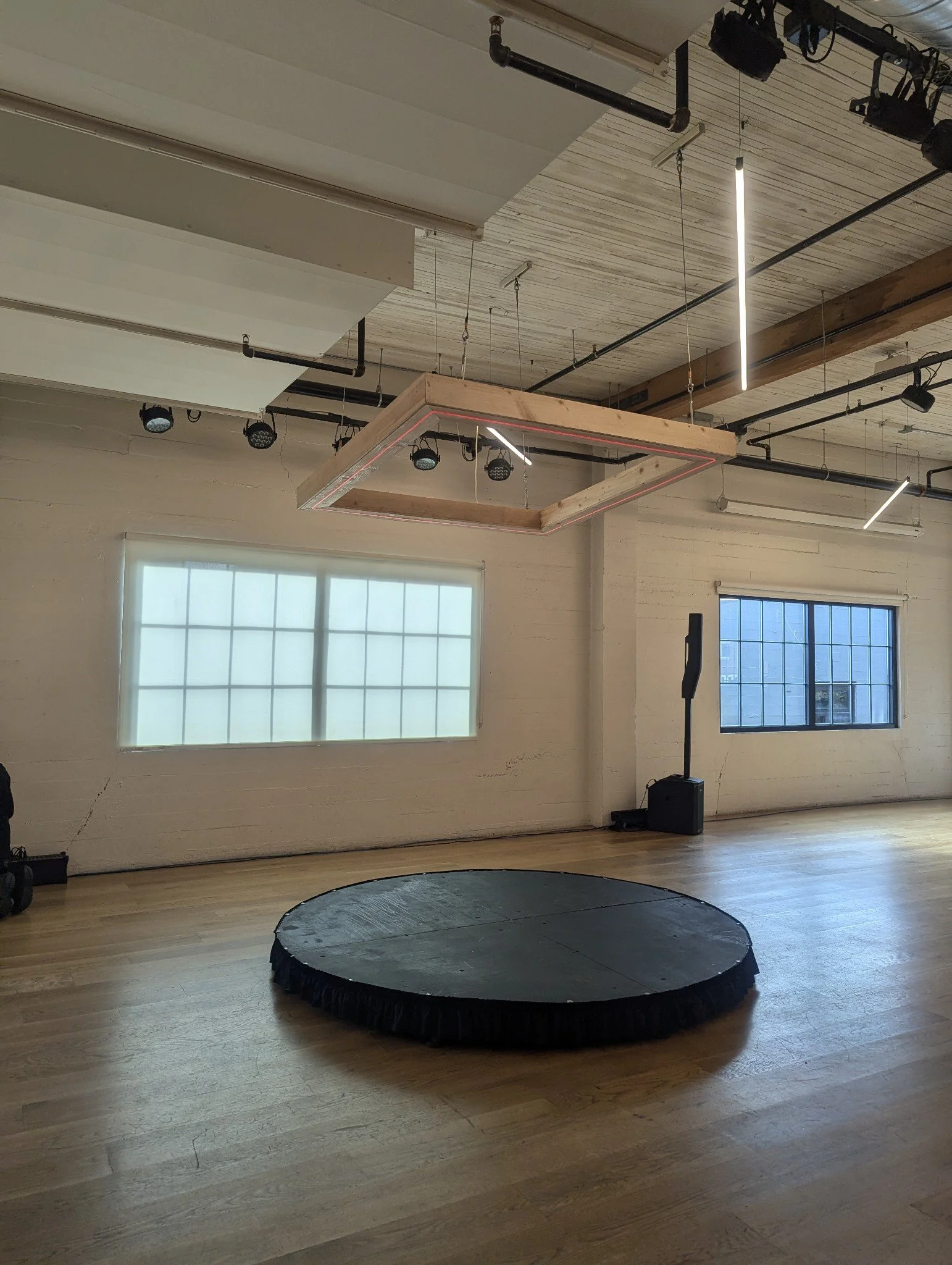 An indoor space with wooden floors and large windows, featuring a small black trampoline with a black safety pad in the foreground, and stage or lighting equipment hanging from the ceiling.