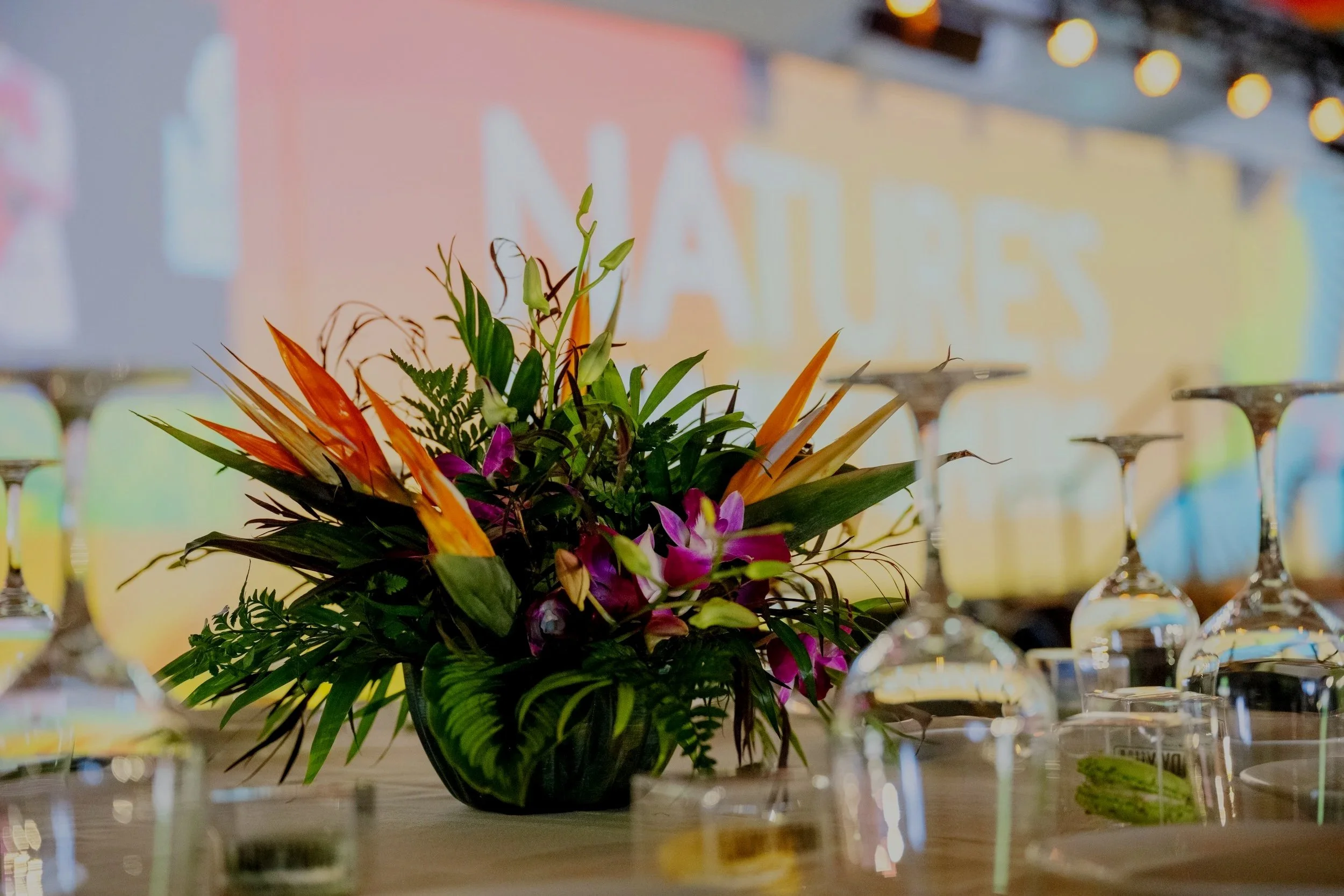 A floral centerpiece with vibrant pink, orange, and green flowers and foliage on a table surrounded by upside-down wine glasses, with a blurred colorful background.