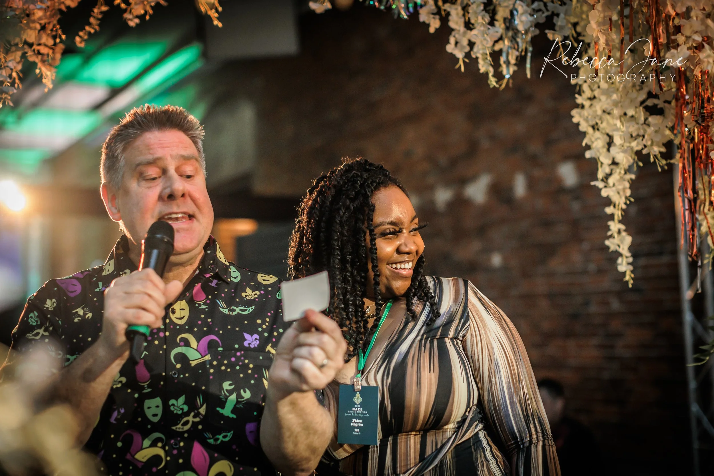 Two people, a man and a woman, stand side by side at an indoor event decorated with hanging flowers and decorations. The man is holding a microphone and reading from a card. The woman is smiling and wearing a striped top with a badge around her neck.