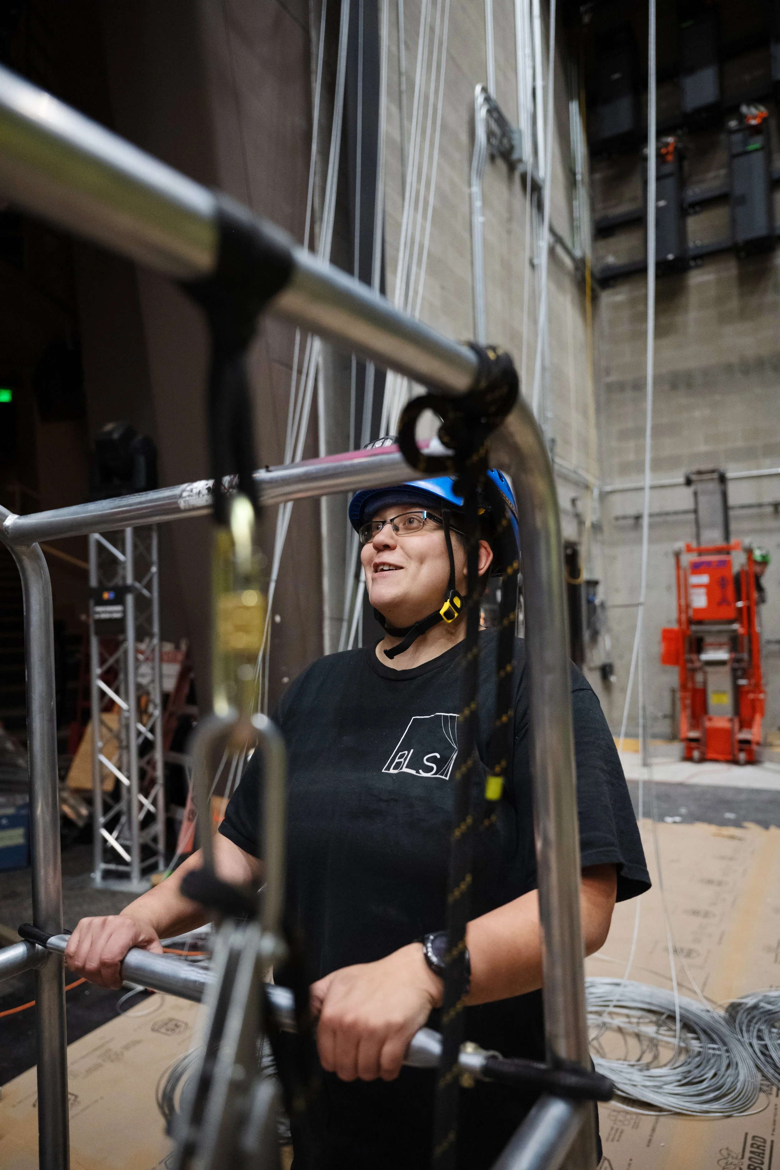 A man wearing glasses and a blue helmet is working with metal pipes and cables in an industrial or construction setting. You can see scaffolding and various equipment in the background.